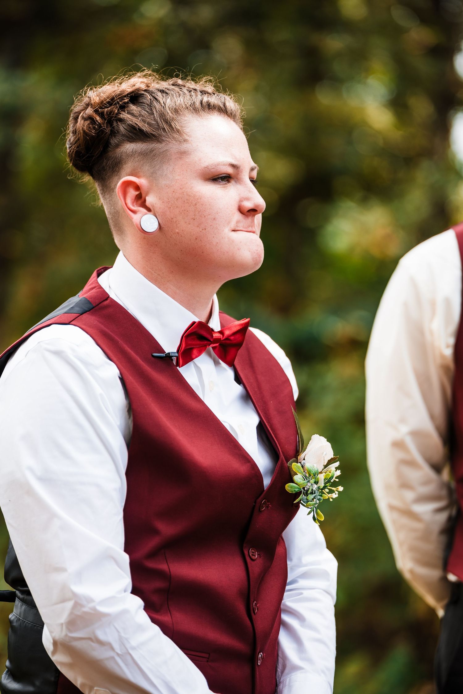 Wedding attendant in burgundy vest and bowtie with boutonniere against natural backdrop.