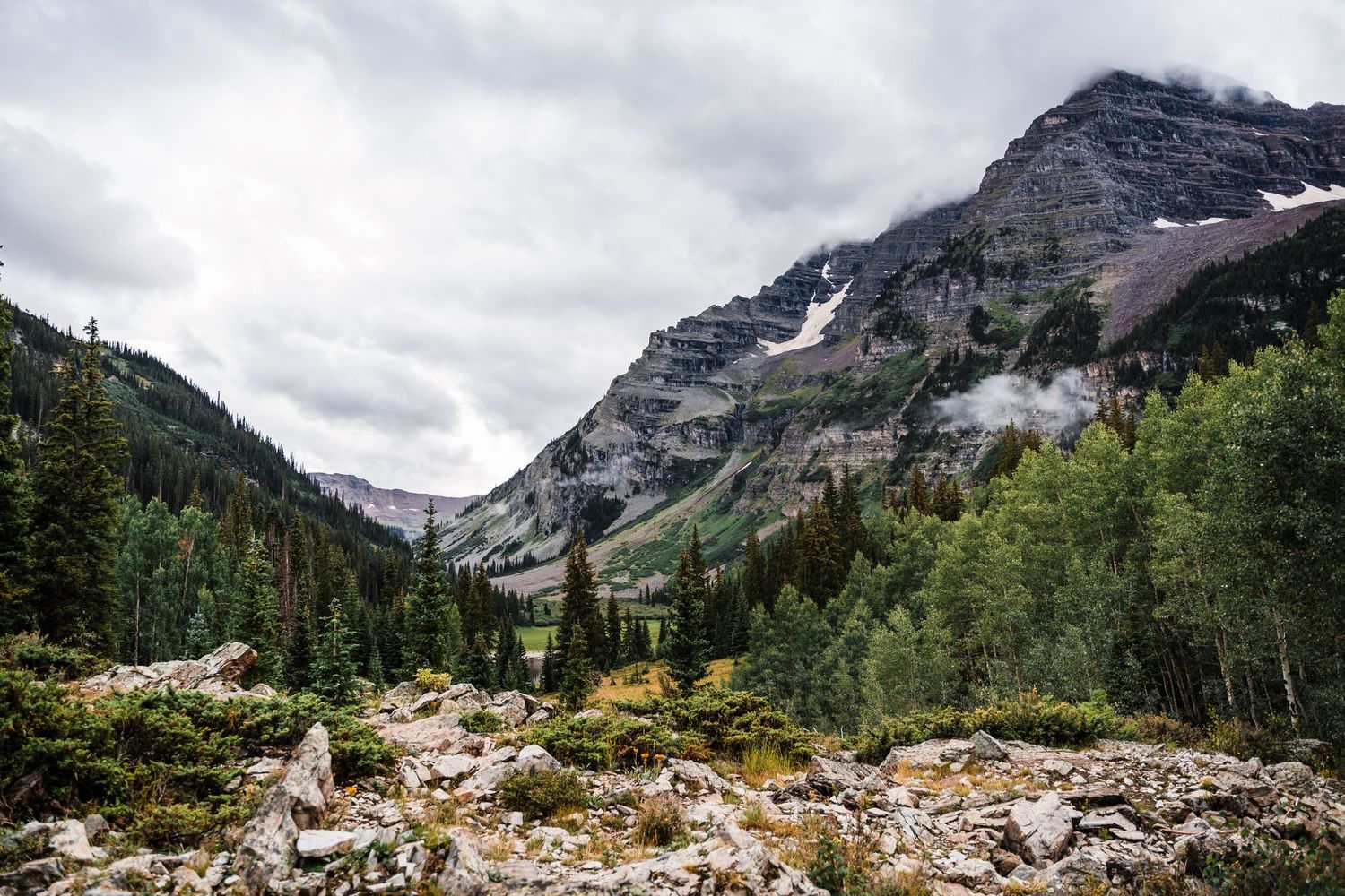 Dramatic mountain valley landscape with snow patches and evergreen forests under cloudy skies.