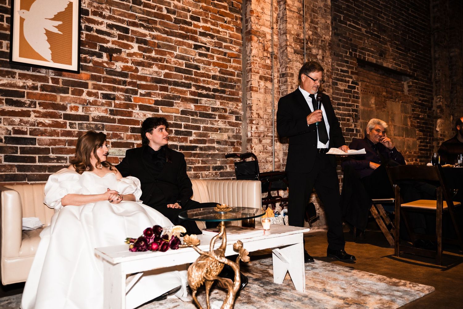 Wedding reception scene with speech being given in rustic brick-walled venue.