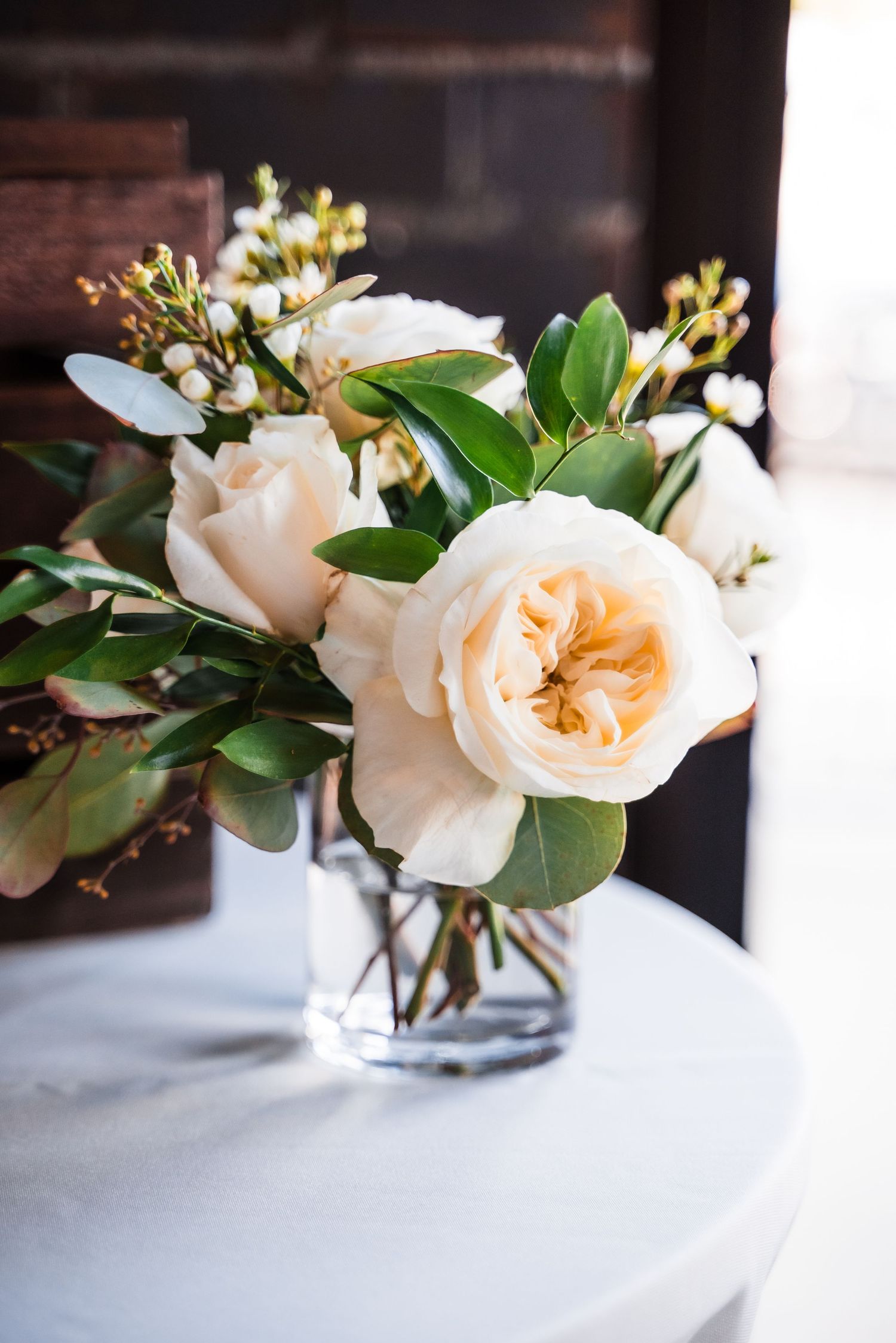Soft peach garden roses and delicate white flowers arranged in a clear glass vase on a white surface.