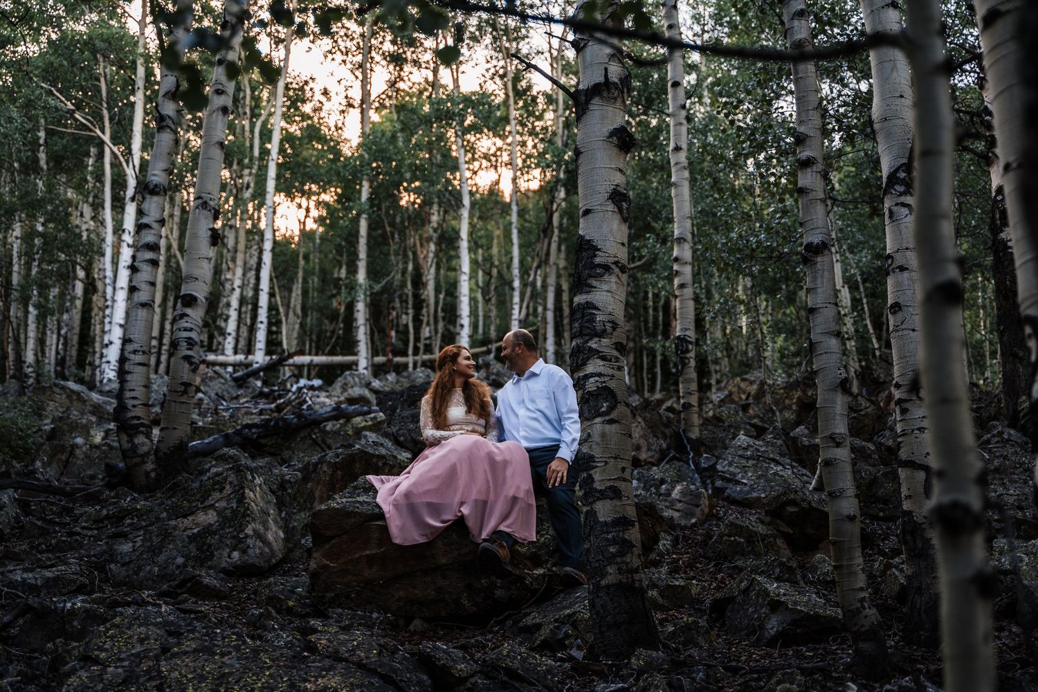 A romantic evening scene among birch trees with a couple sitting on fallen logs at sunset.