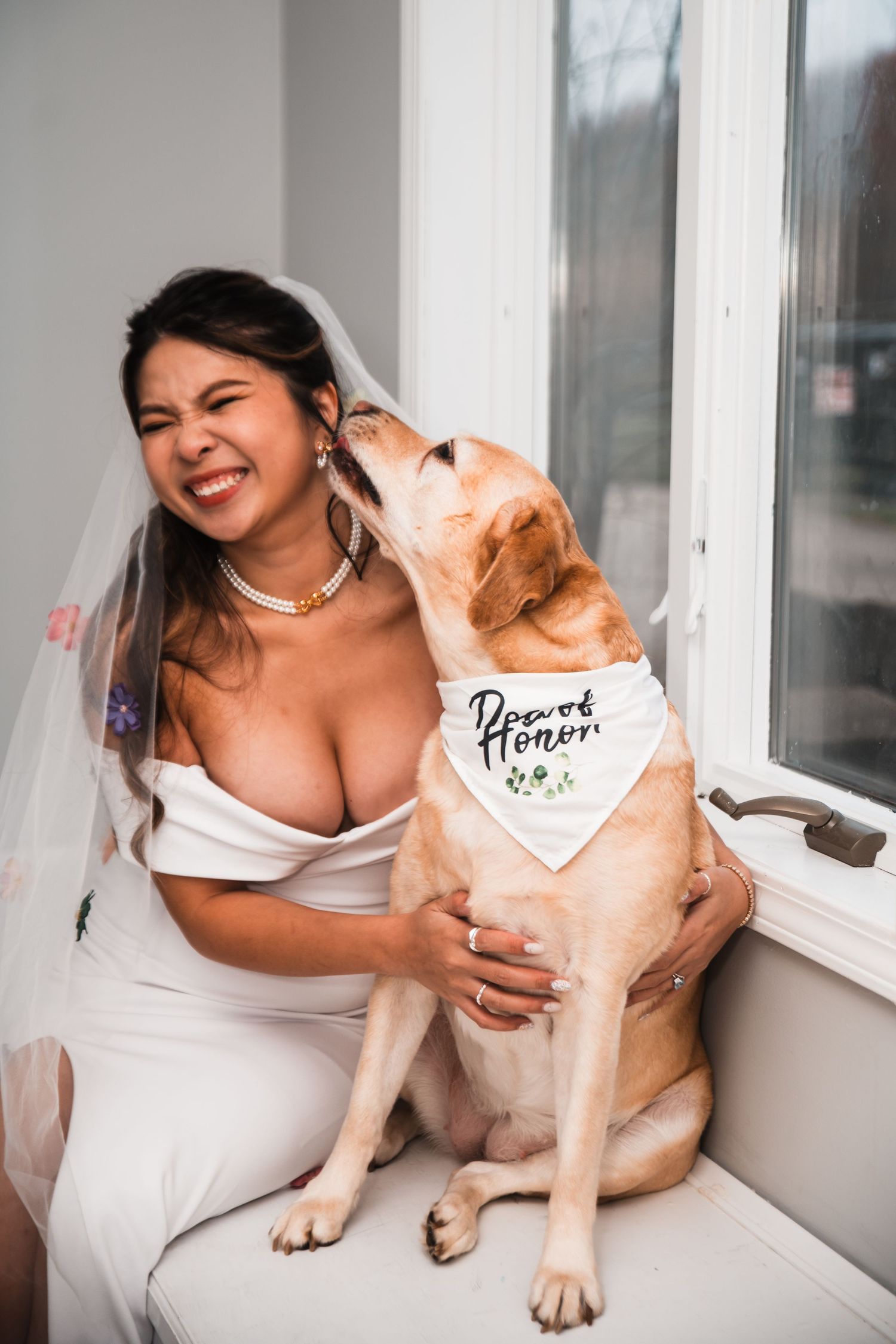 A bride in white dress sits with her dog who wears a wedding-themed bandana.