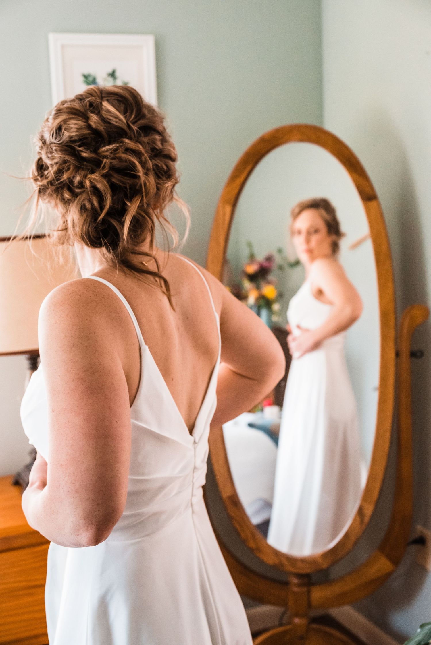 A bride in a white dress looking at her reflection in a vintage oval mirror.