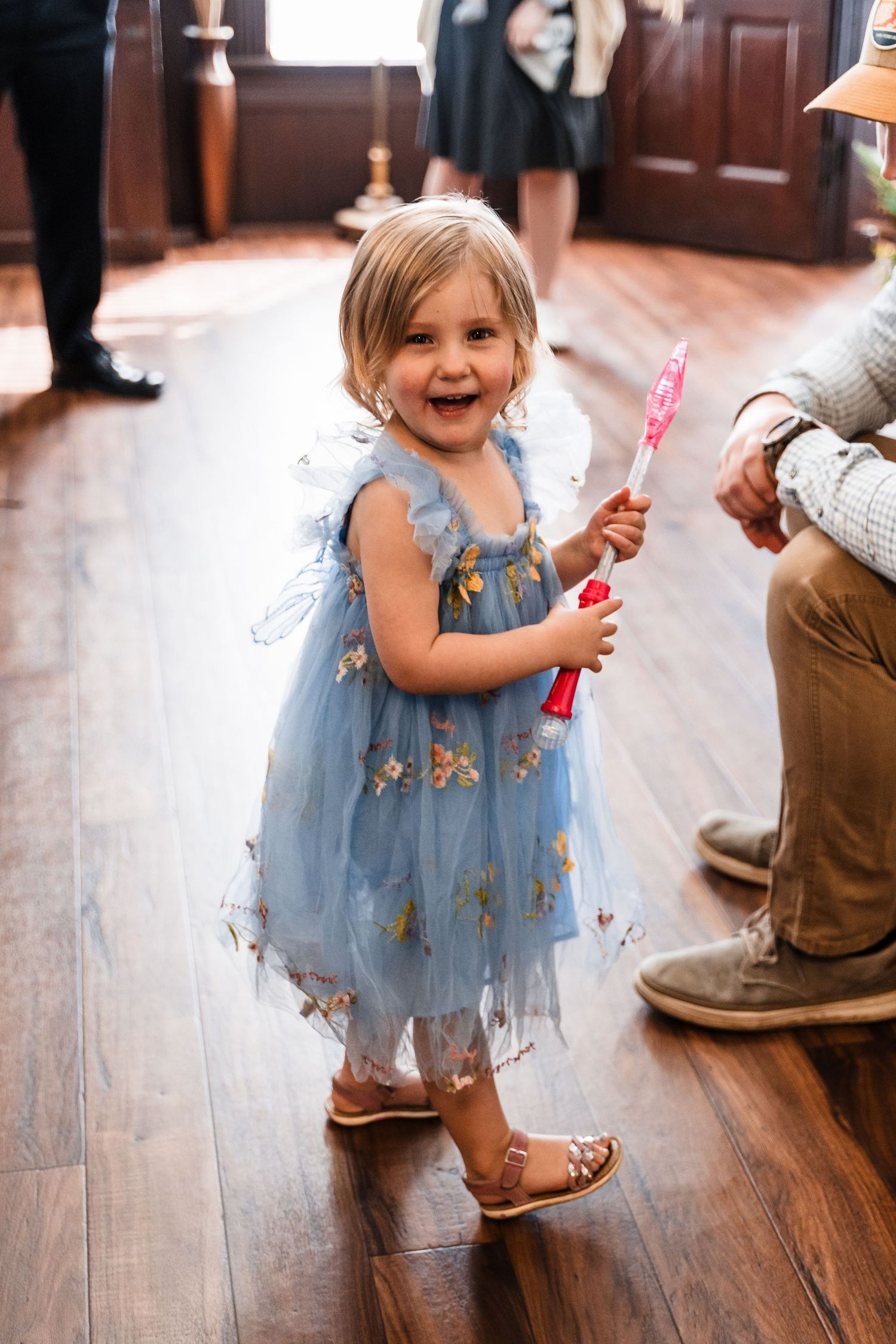 Smiling toddler girl in blue floral tulle dress holding pink wand at wedding celebration.