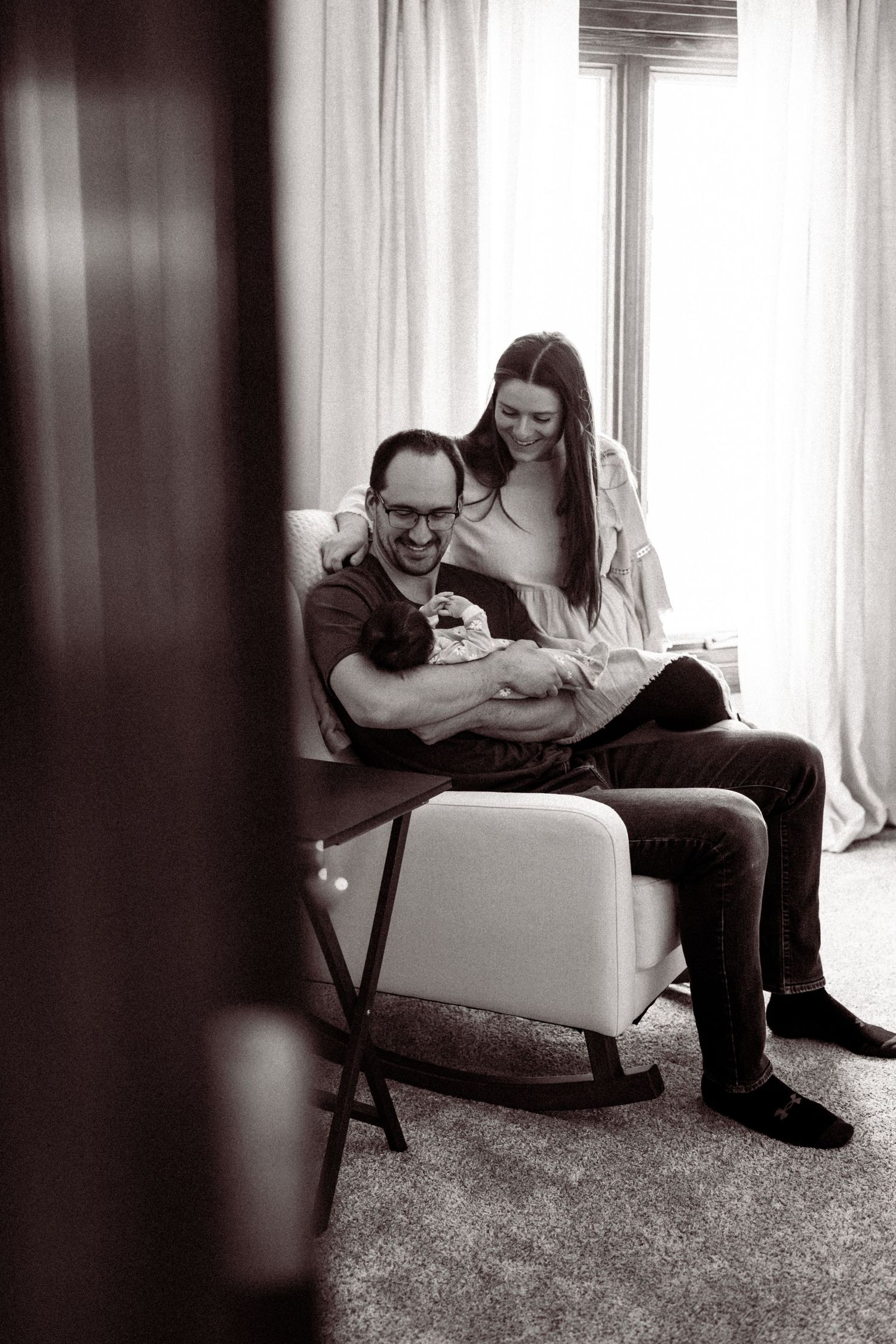 A black and white photograph of a couple sitting together with a newborn baby by a window with sheer curtains.