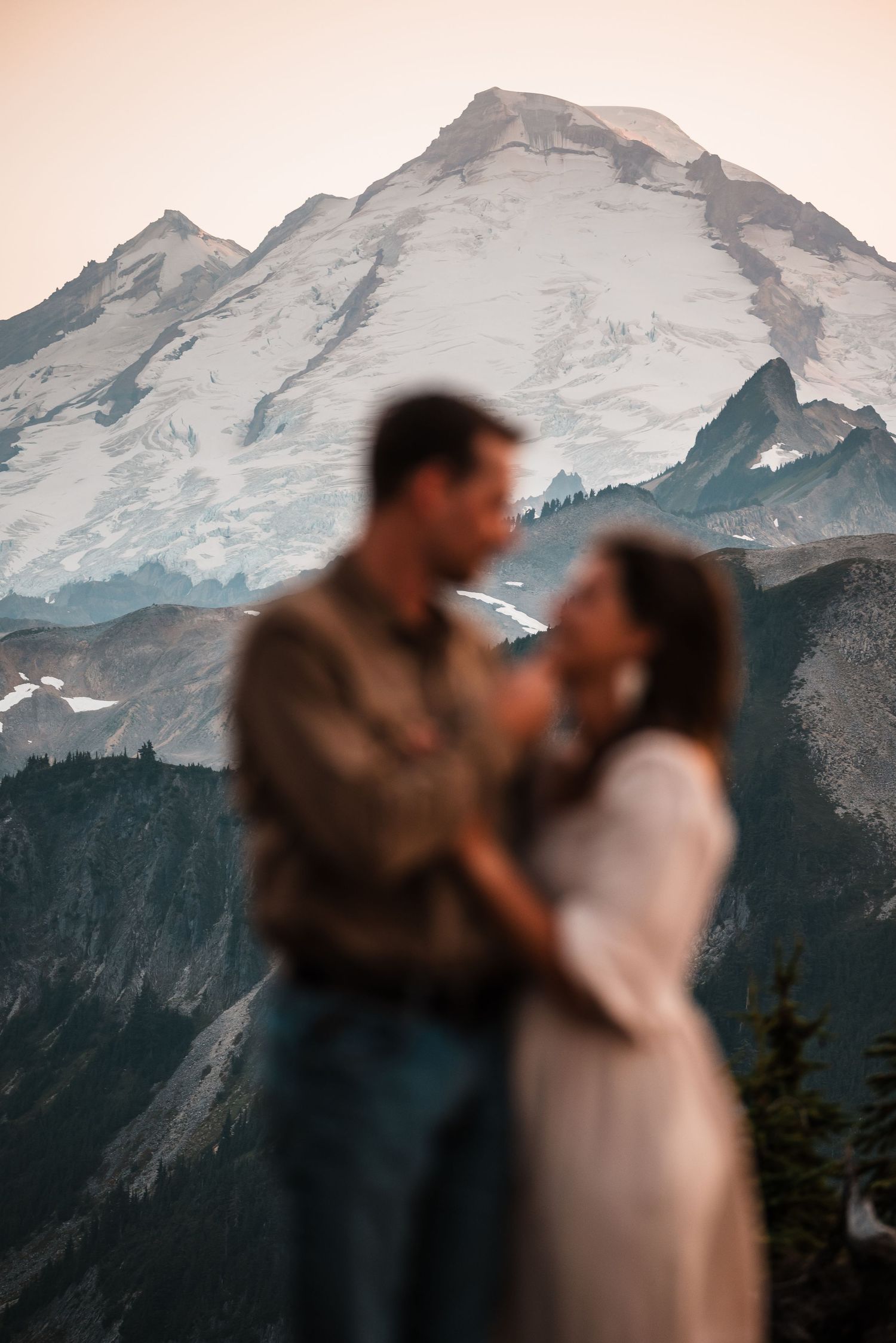 A couple embraces in silhouette against a dramatic mountain backdrop with snow-capped Mount Rainier peaks at sunset.