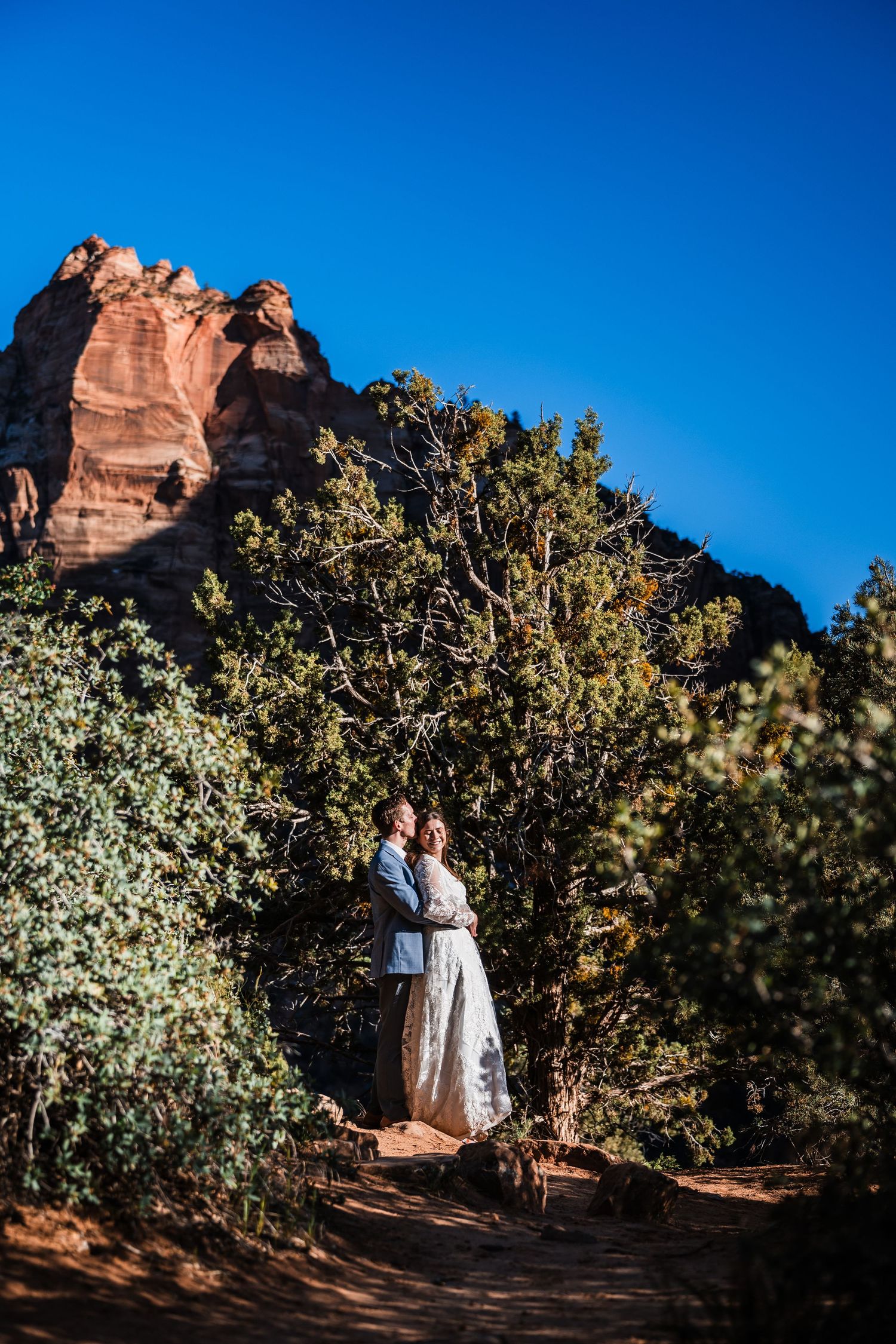 Wedding couple embraces on desert trail surrounded by lush trees and red rock formations under blue sky.