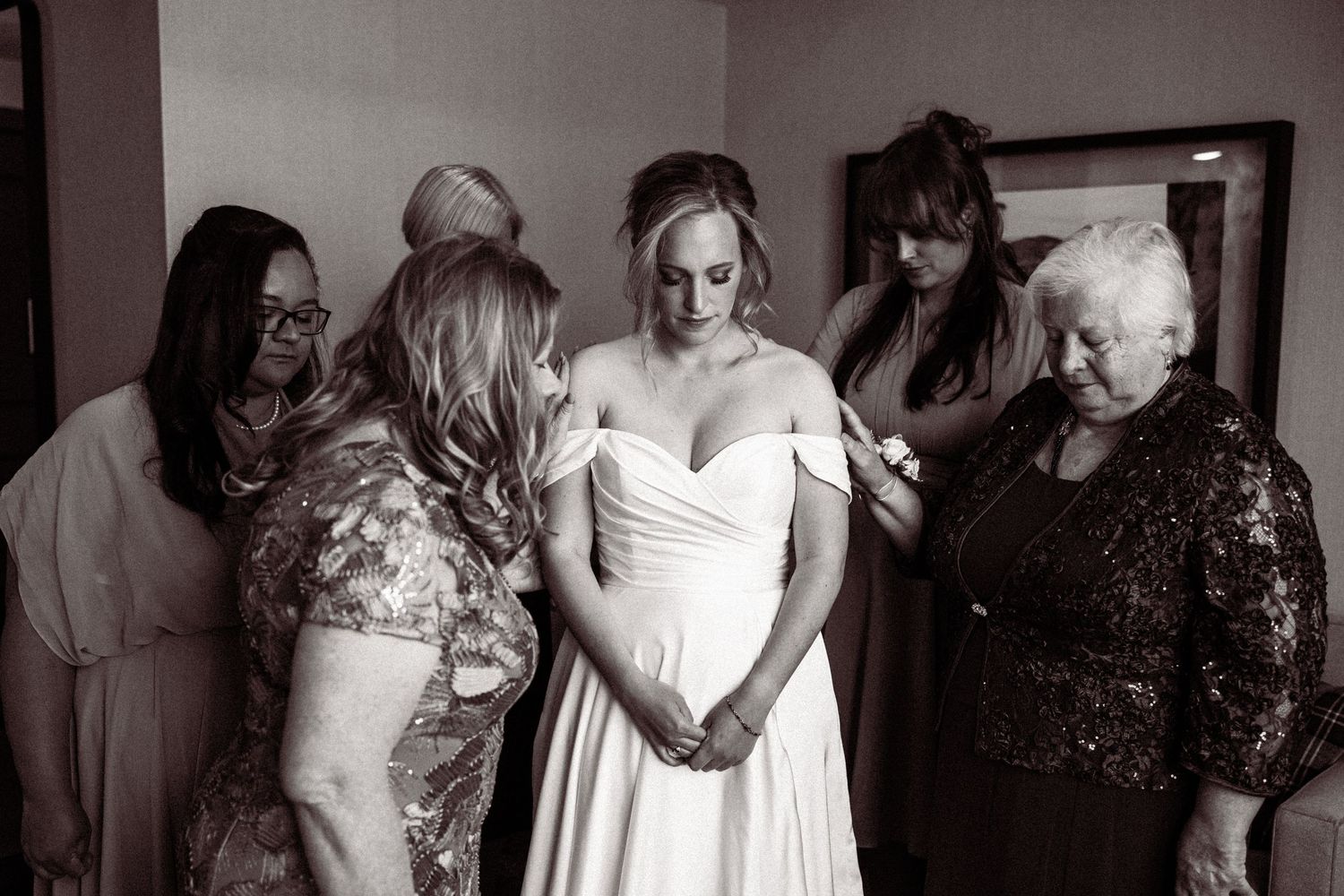 Bride in white wedding gown surrounded by family members bowing heads in prayer before ceremony, black and white photo.