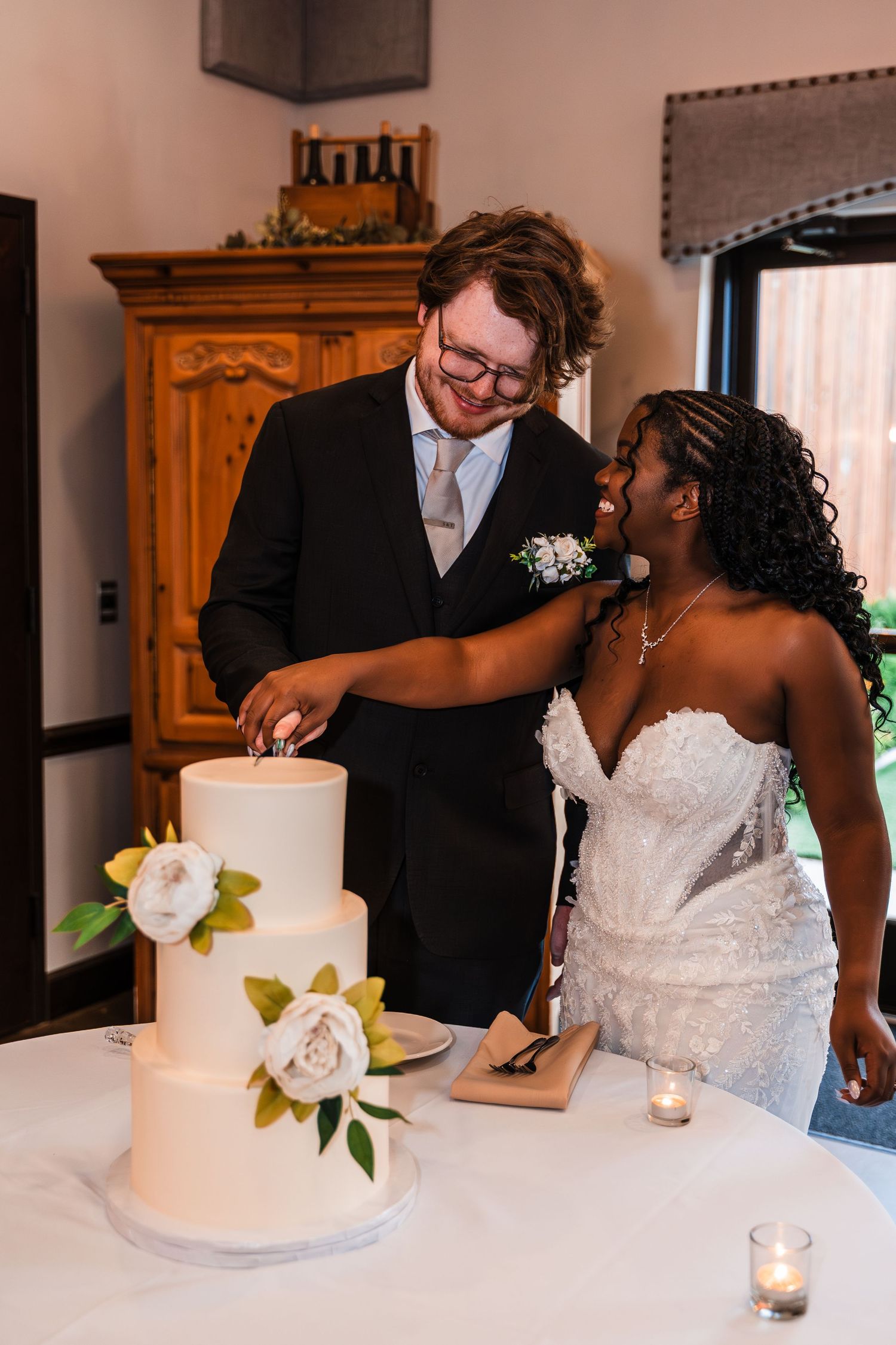 Bride and groom laughing while cutting a three-tier white wedding cake decorated with flowers.