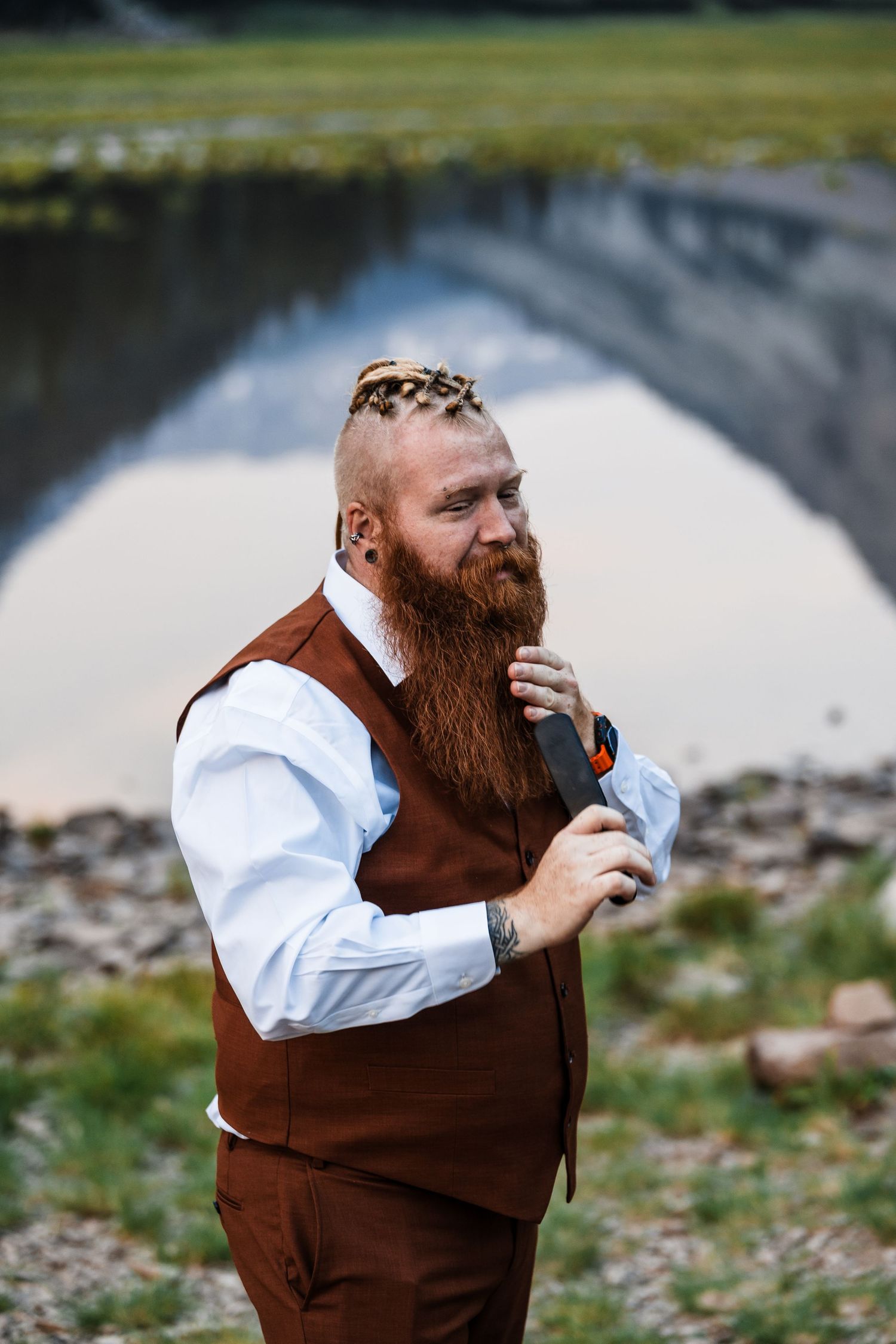 Bearded groom in brown vest adjusts tie near reflective pond at outdoor wedding.