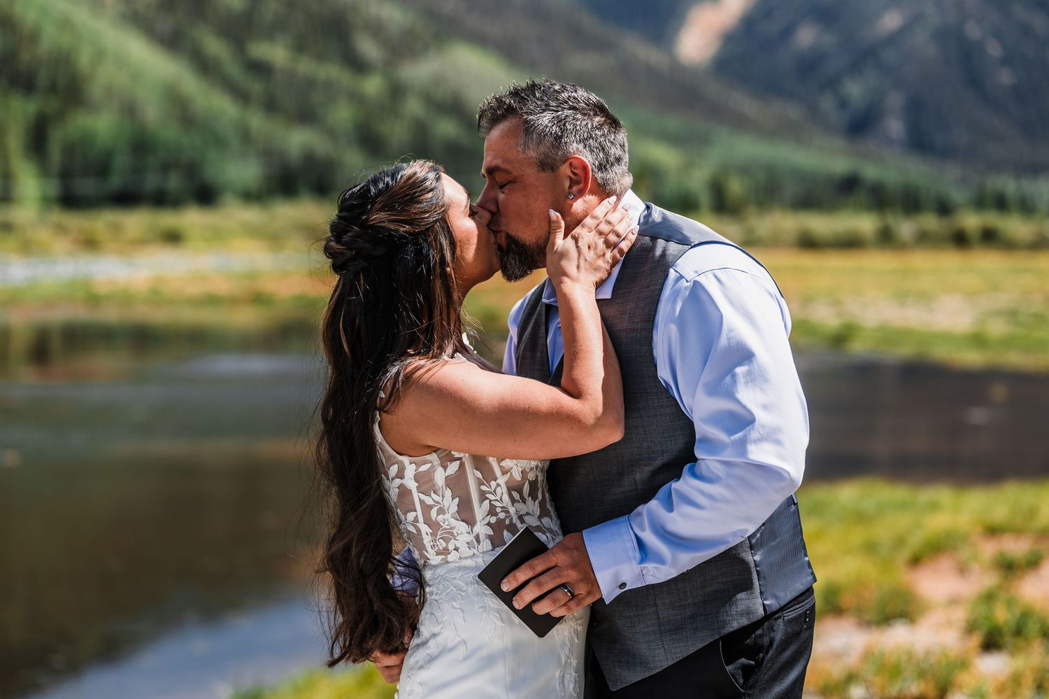 A couple shares a romantic kiss at their outdoor wedding ceremony with a scenic mountain river backdrop on a sunny day.