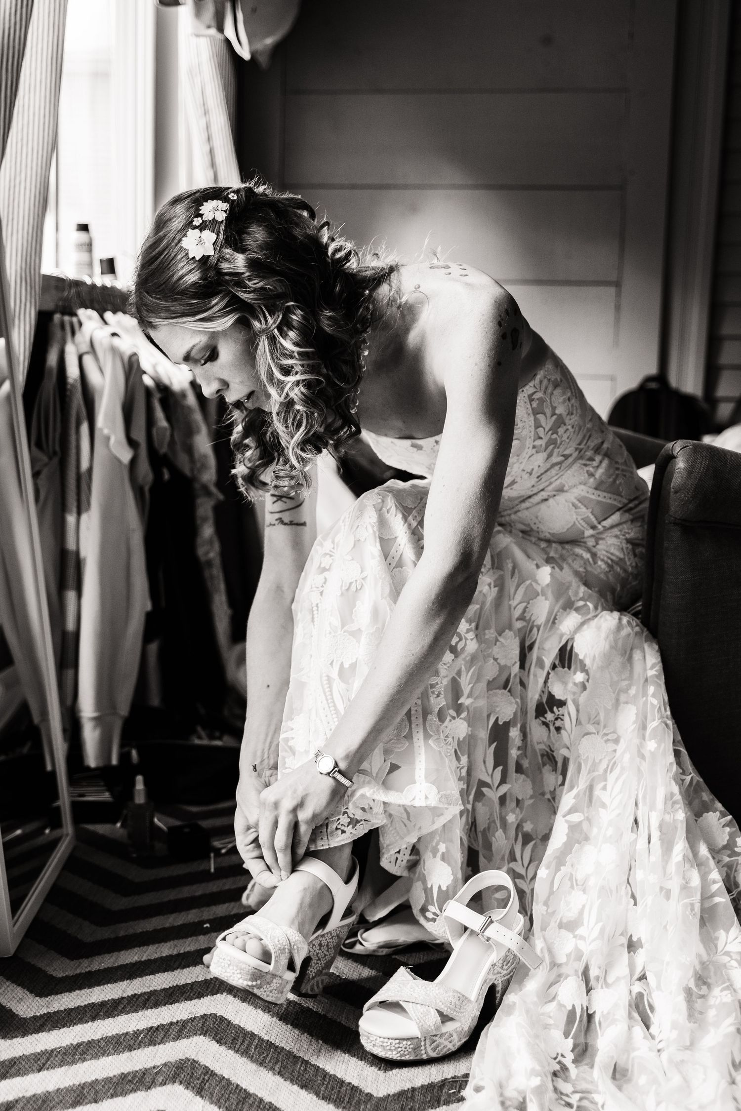 Black and white photo of someone in a lace wedding dress fastening white sandals while seated on a striped carpet.