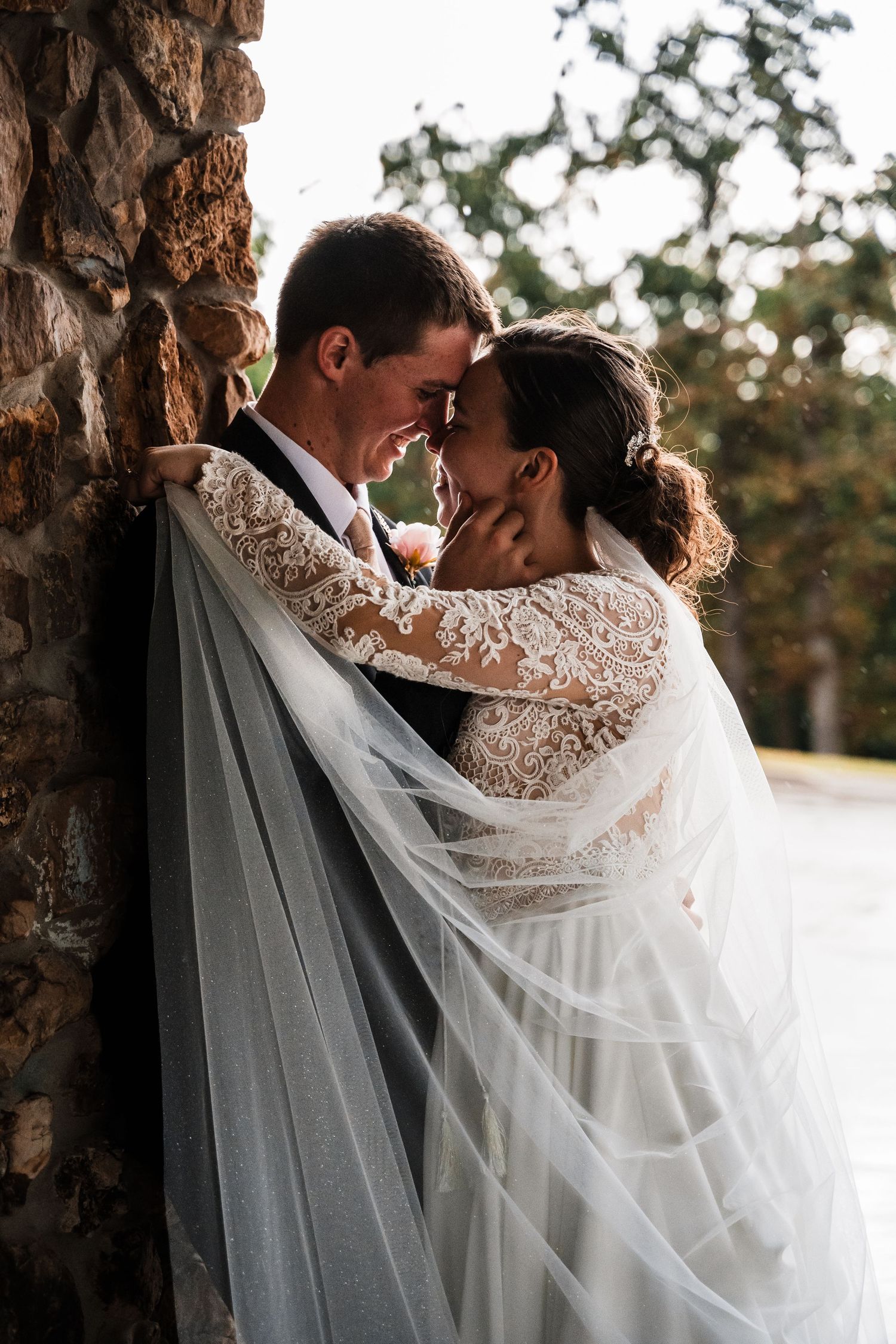 A romantic wedding couple embraces near a tree during golden hour, with the bride's lace veil flowing in the breeze.