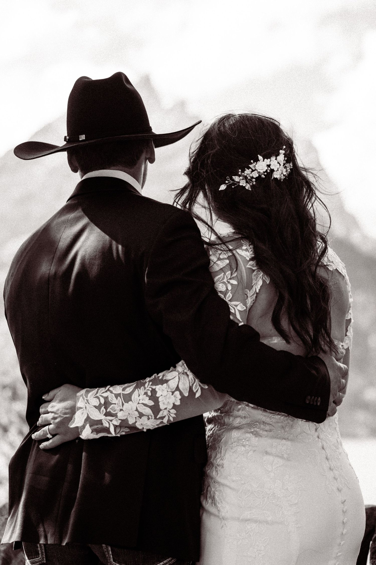 Black and white wedding photo of a couple embracing with mountains in background, bride in white and groom in cowboy hat.
