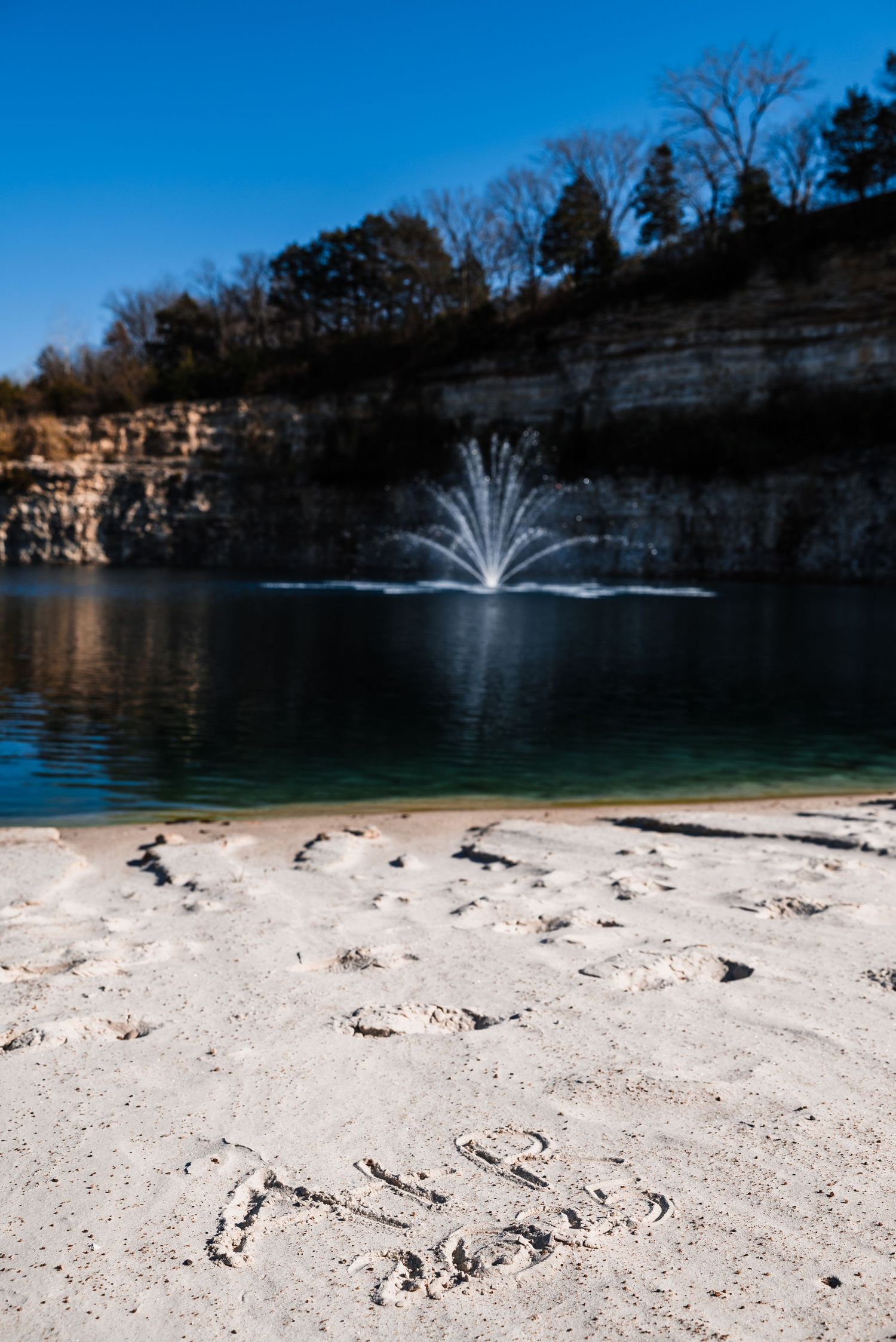 A water fountain sprays in a quarry lake with limestone cliffs and sandy beach in the foreground.