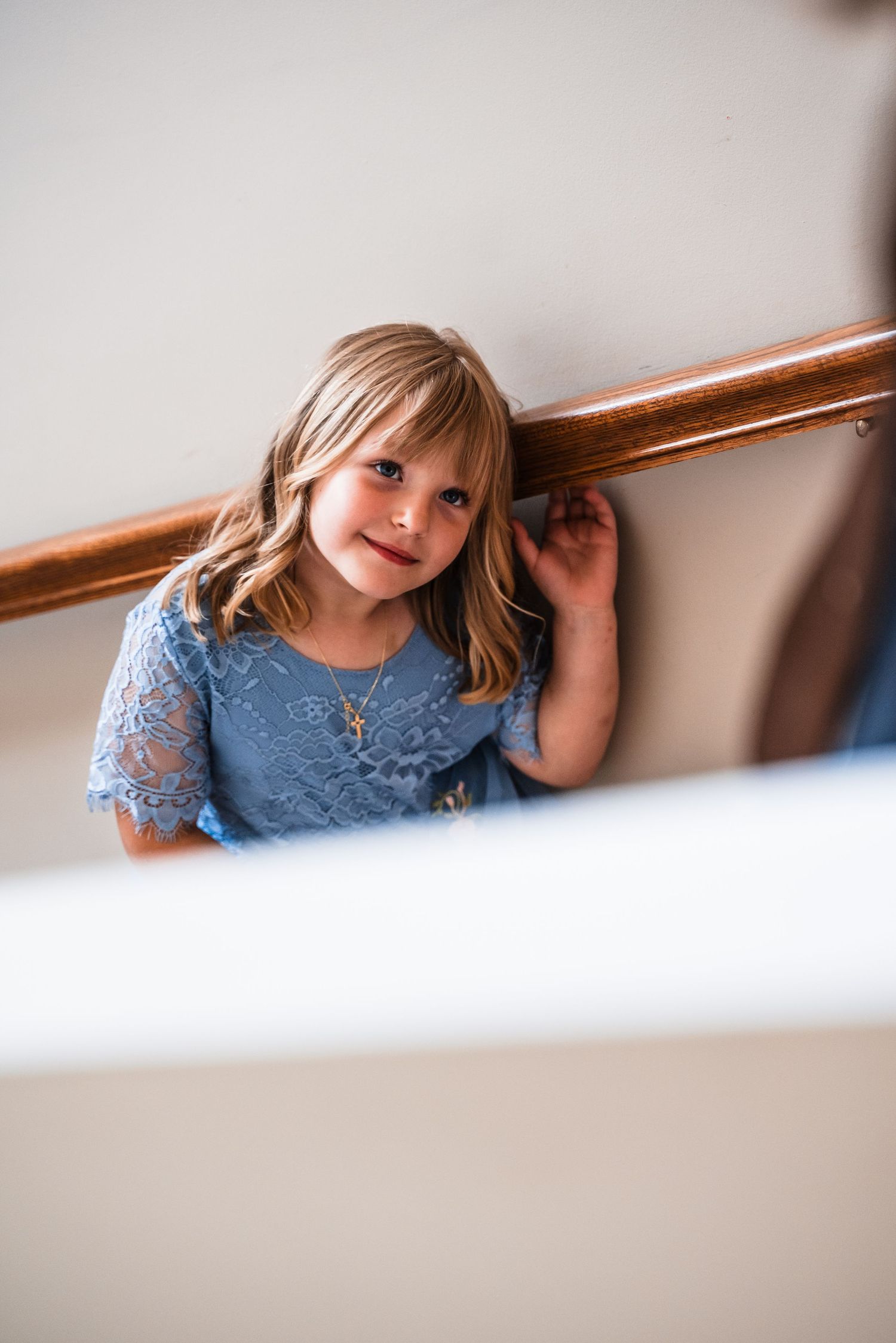 A young child in a blue lace dress poses by a wooden railing.