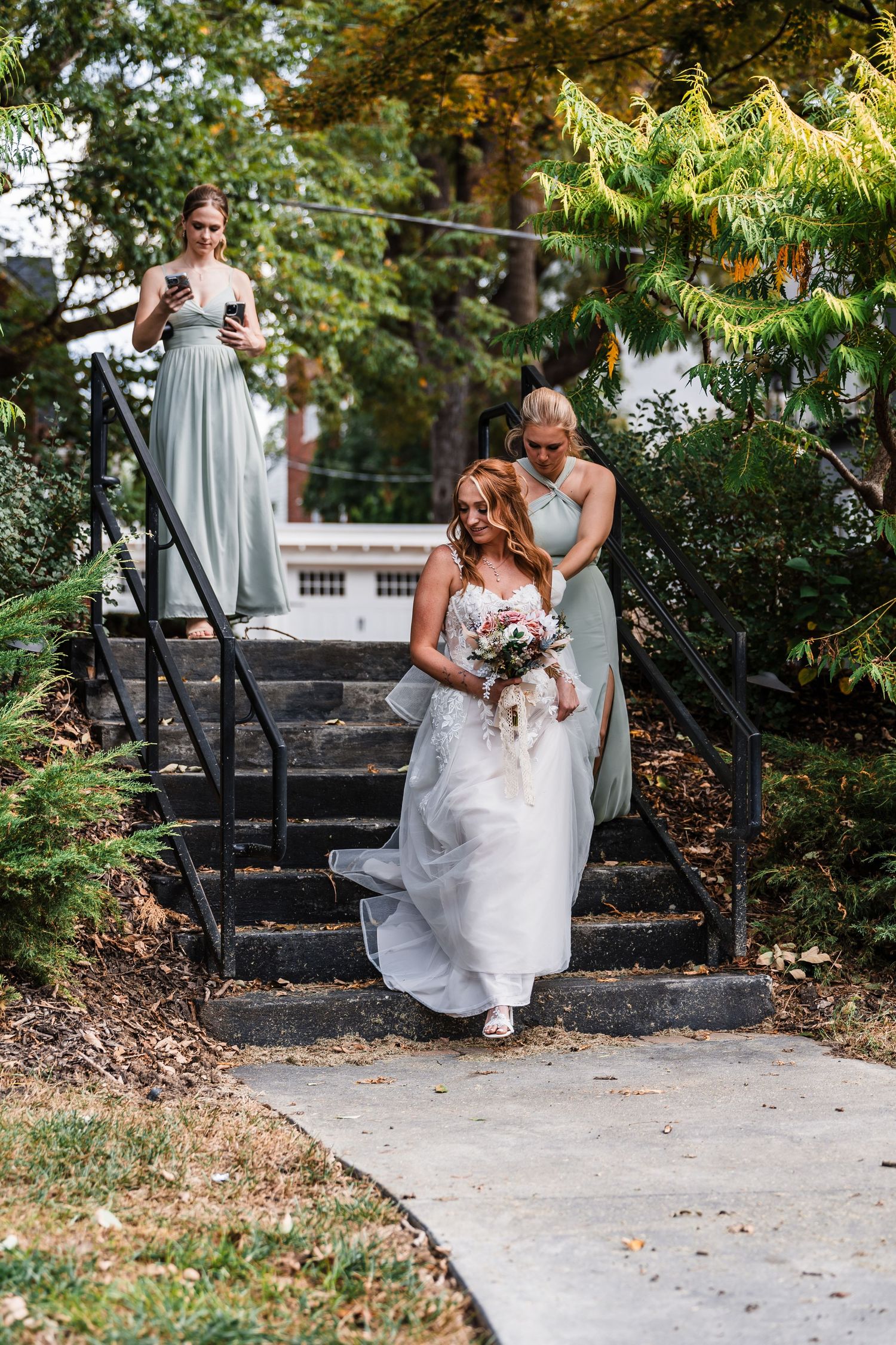 Bride in flowing white gown descends outdoor stone steps surrounded by lush greenery.