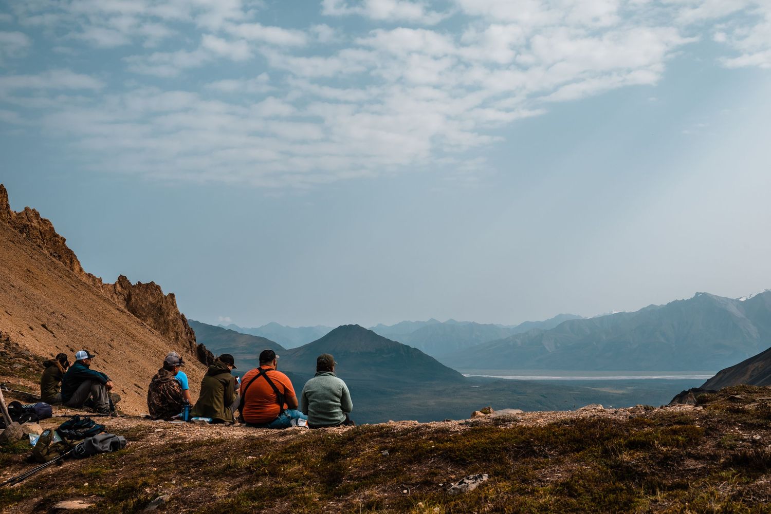Hikers rest on a mountainside overlooking a scenic valley with snow-capped peaks in the distance.