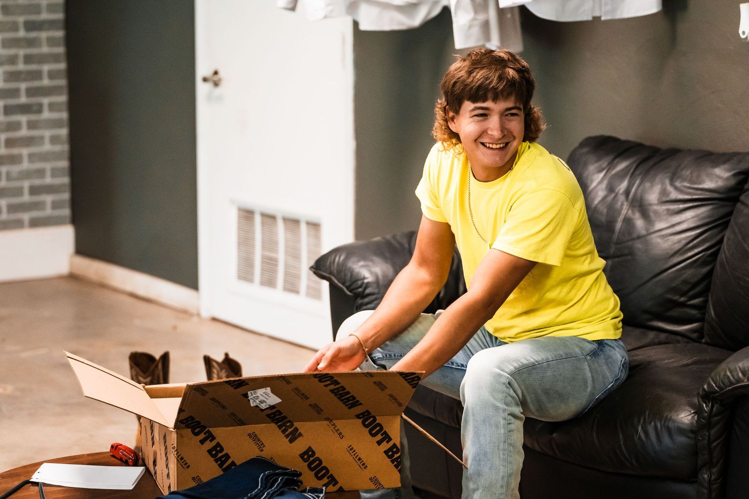 A young person sits on a leather couch while opening a cardboard package with excitement.