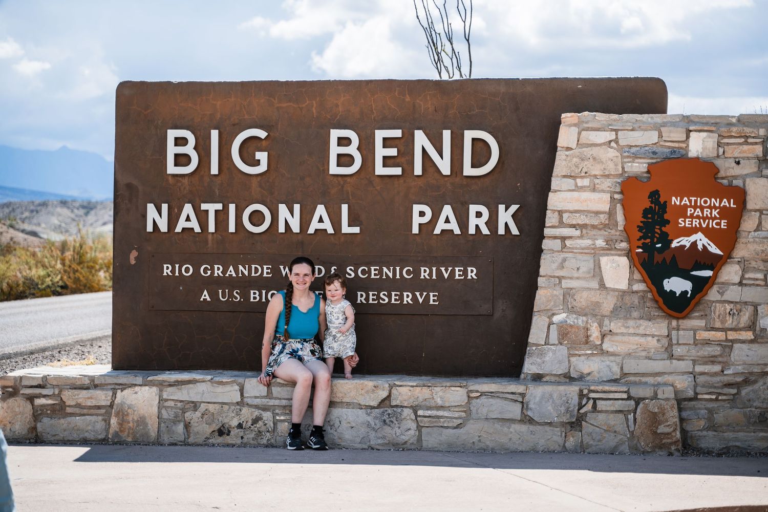 Visitors pose at the entrance sign of Big Bend National Park with mountain landscapes in the background.