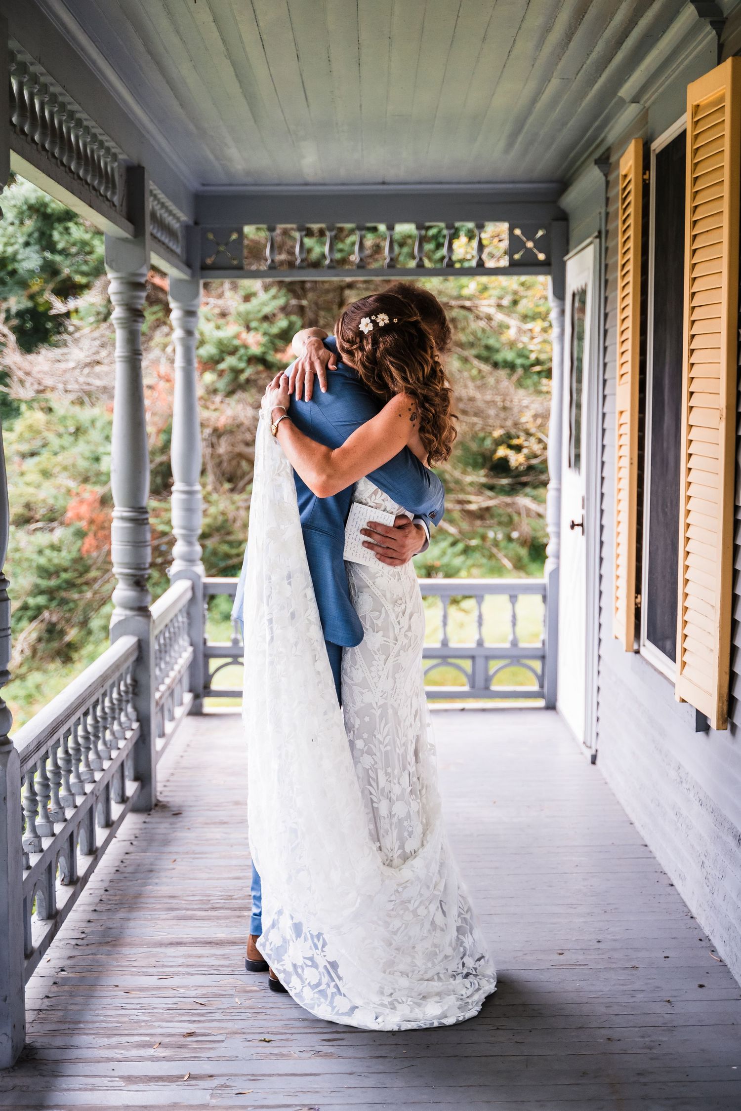Newlyweds embrace on elegant Victorian porch surrounded by natural foliage.