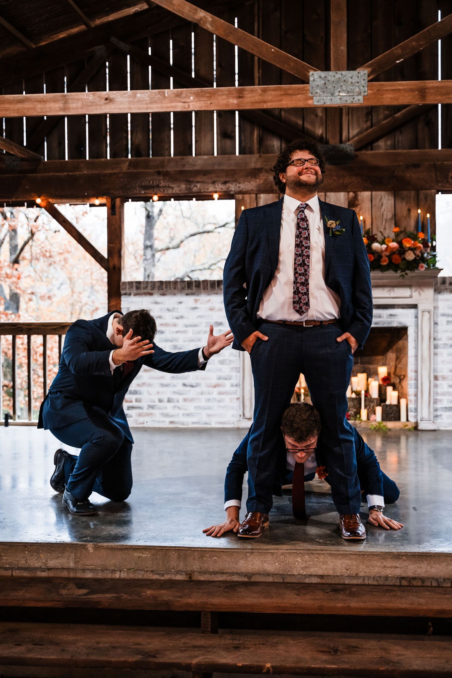 Playful groomsmen striking fun poses on wooden stage at barn venue.