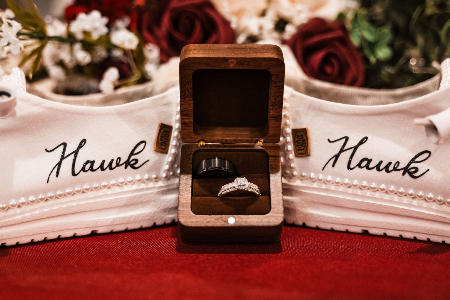 Wedding ring in wooden box displayed between custom shoes with 'Hawk' embroidery.