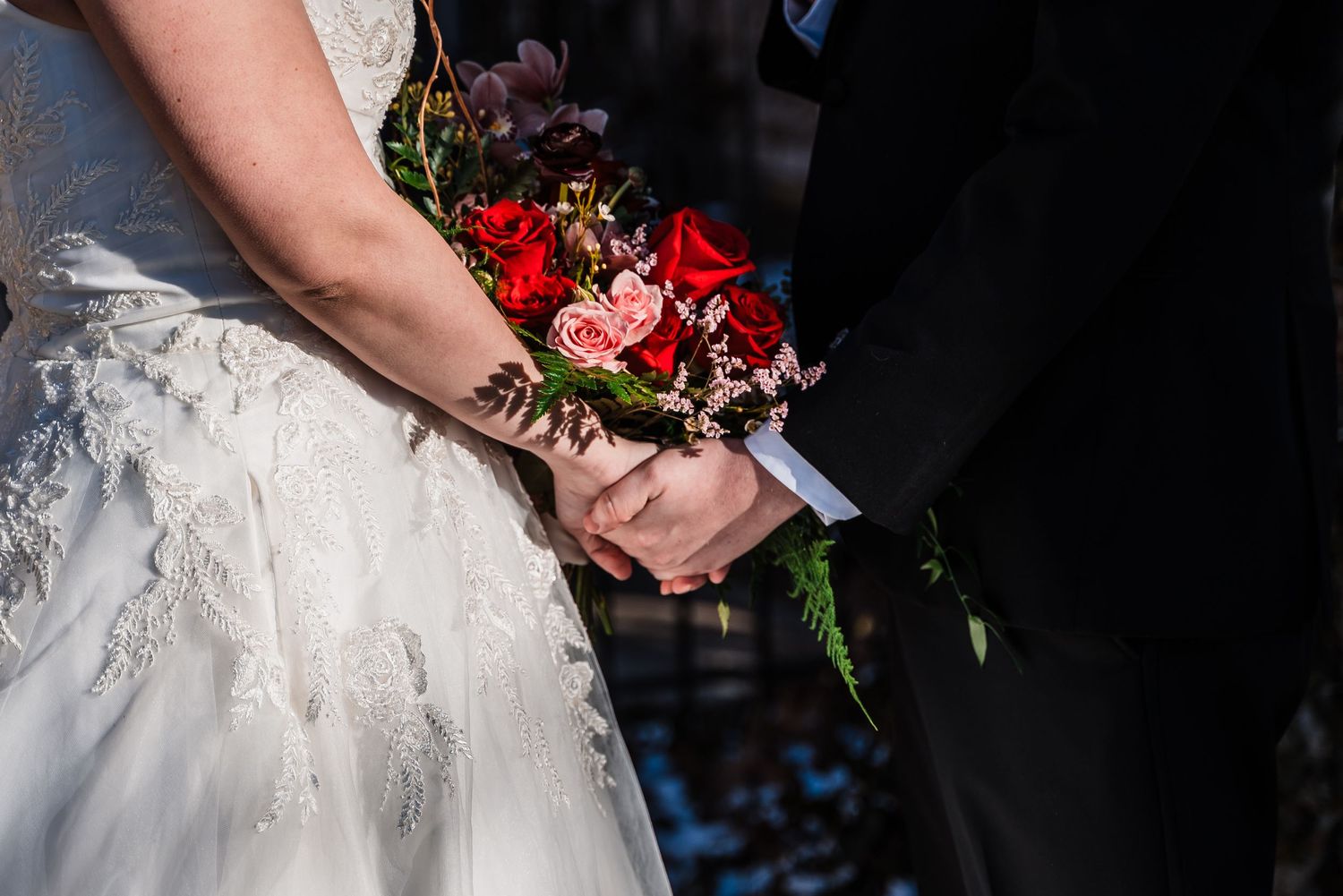 Close-up of bride and groom's hands holding red wedding bouquet against formal attire.