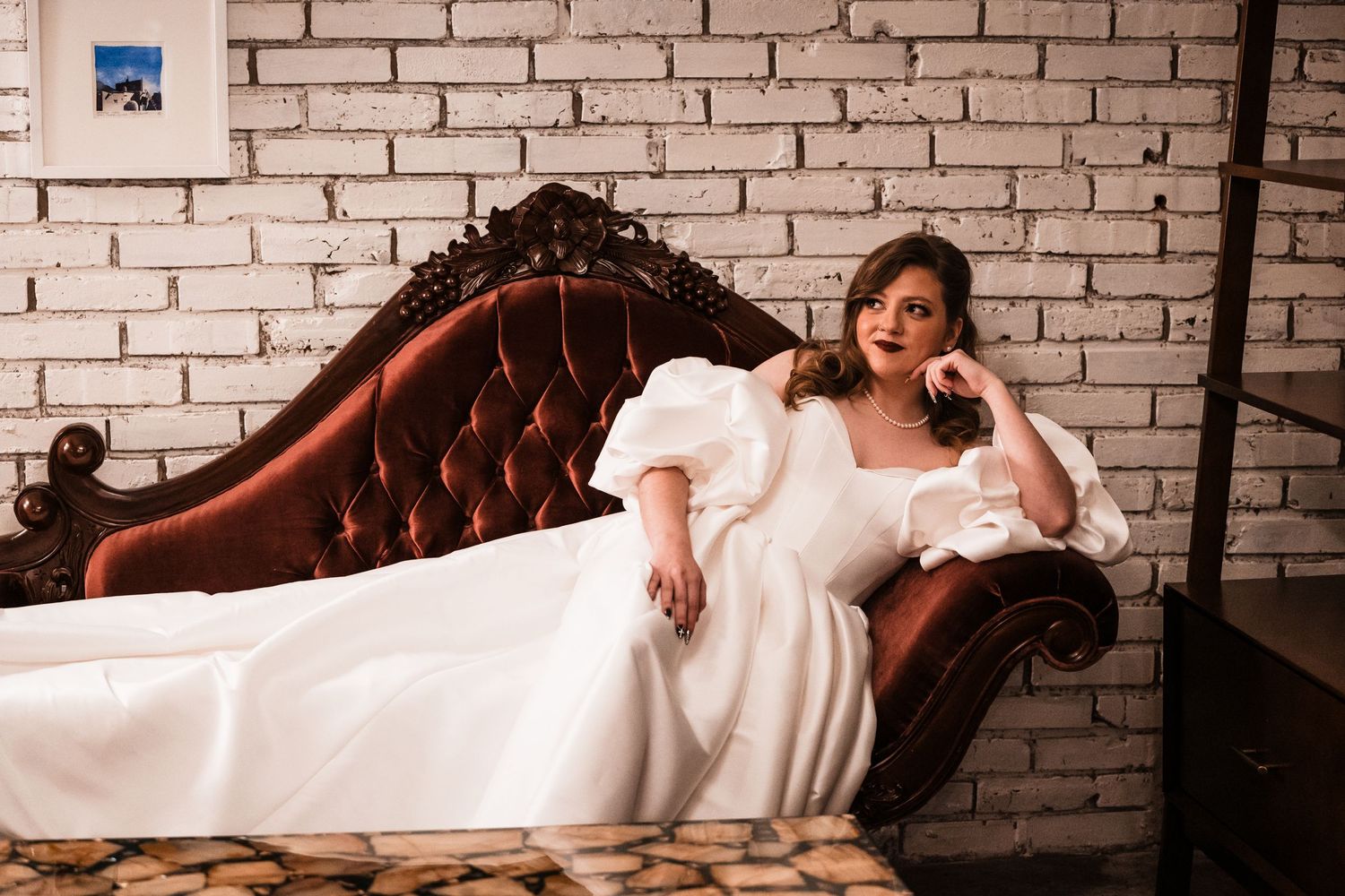 A bride reclines on an elegant tufted brown leather couch against a brick wall in a vintage-inspired portrait.