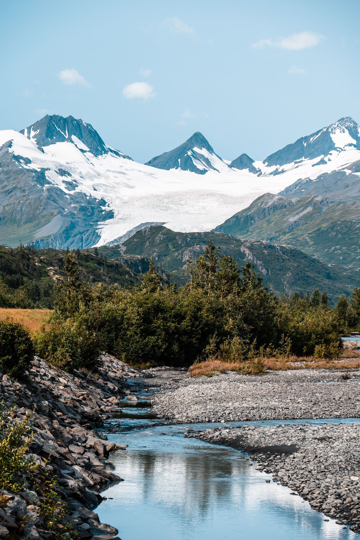 Snow-capped mountain peaks tower above a pristine stream flowing through a rocky valley.