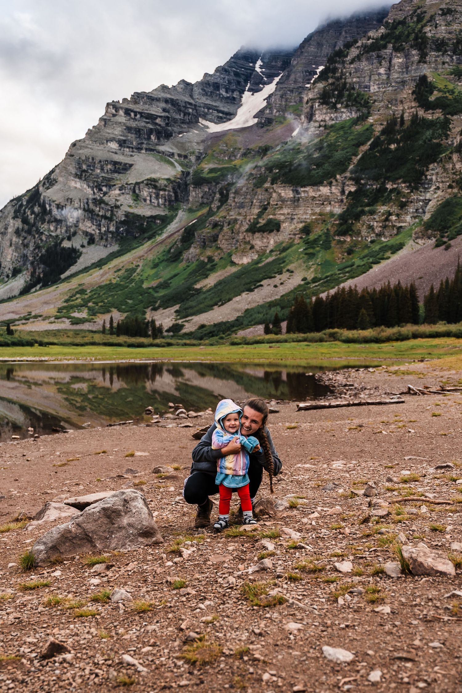 Hikers resting by mountain lake with towering rock face and snow patches in background.