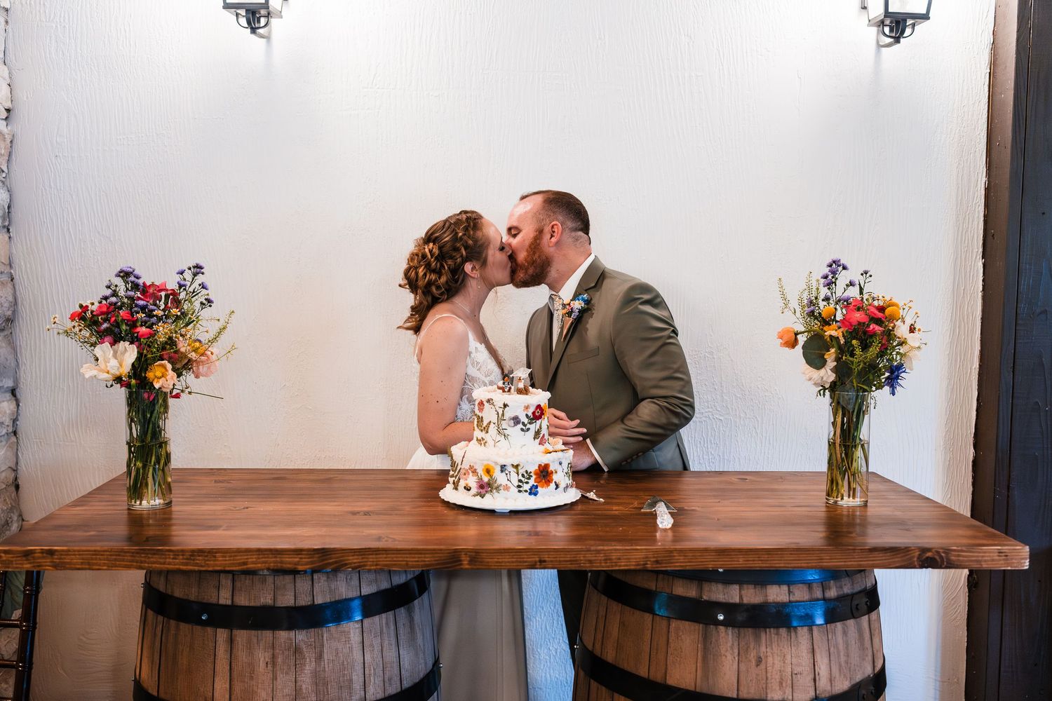 Bride and groom share first kiss as married couple behind wedding cake on barrel table.