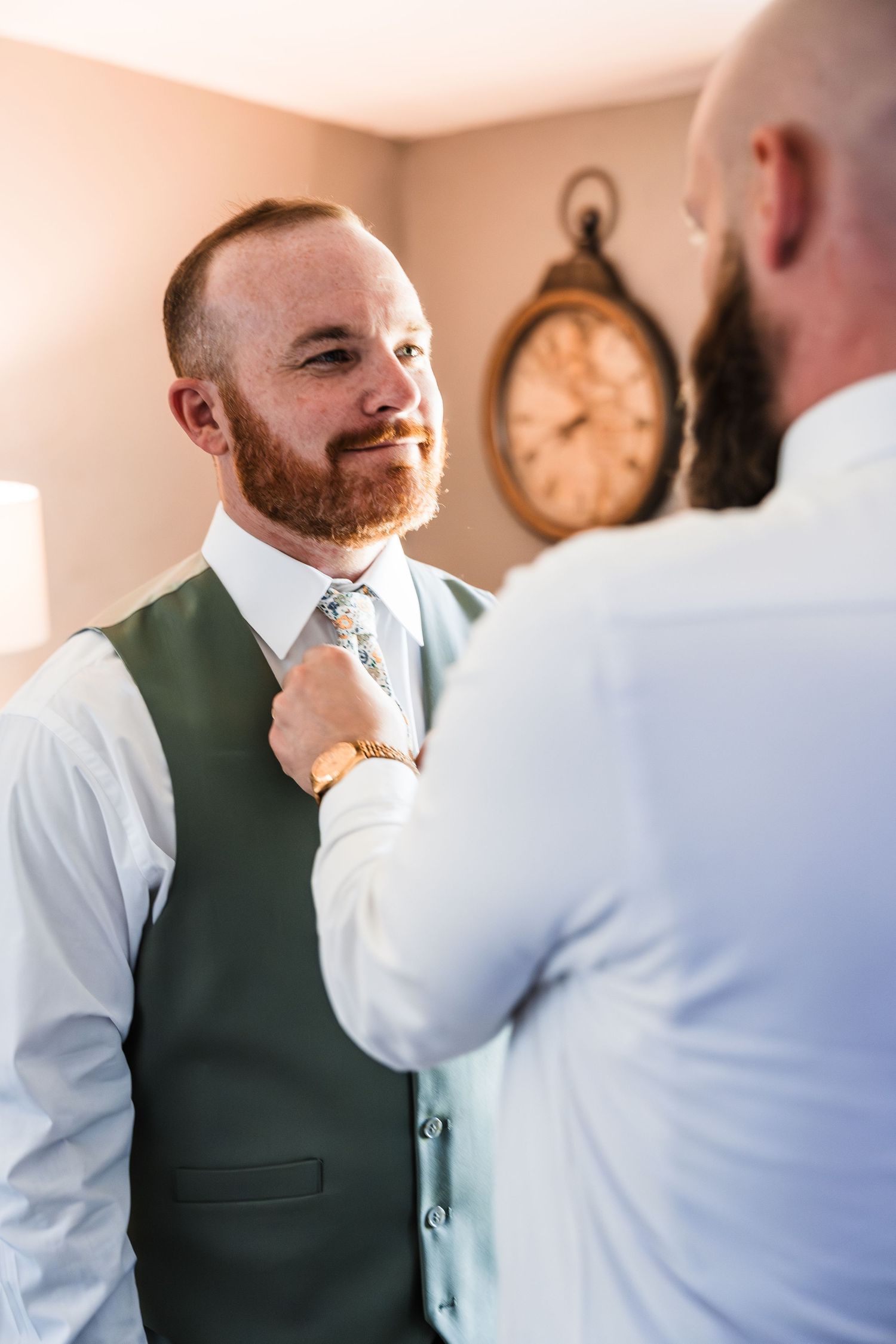 Groom in green vest gets tie adjusted by groomsman before wedding ceremony.