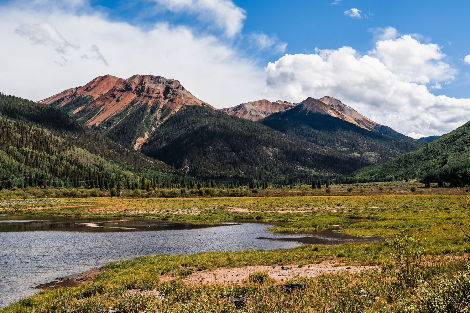 Scenic mountain landscape with red peaks, evergreen forest, and a tranquil pond under a partly cloudy sky in Colorado.
