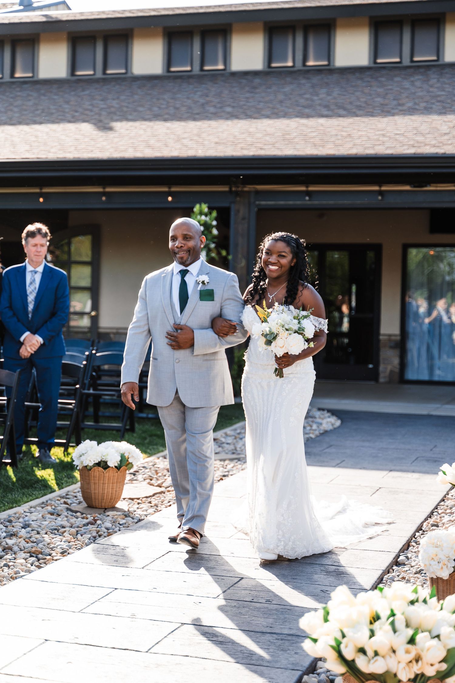 Smiling bride in white gown walks down outdoor aisle with escort at sunny wedding ceremony.