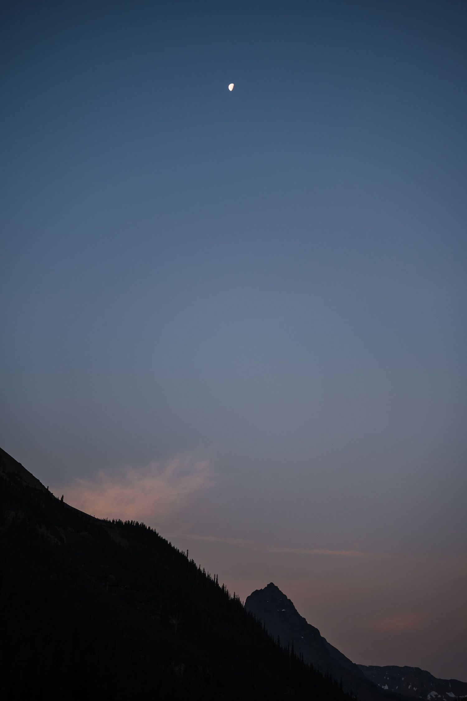 Crescent moon glowing in blue dusk sky above mountain silhouette at twilight.