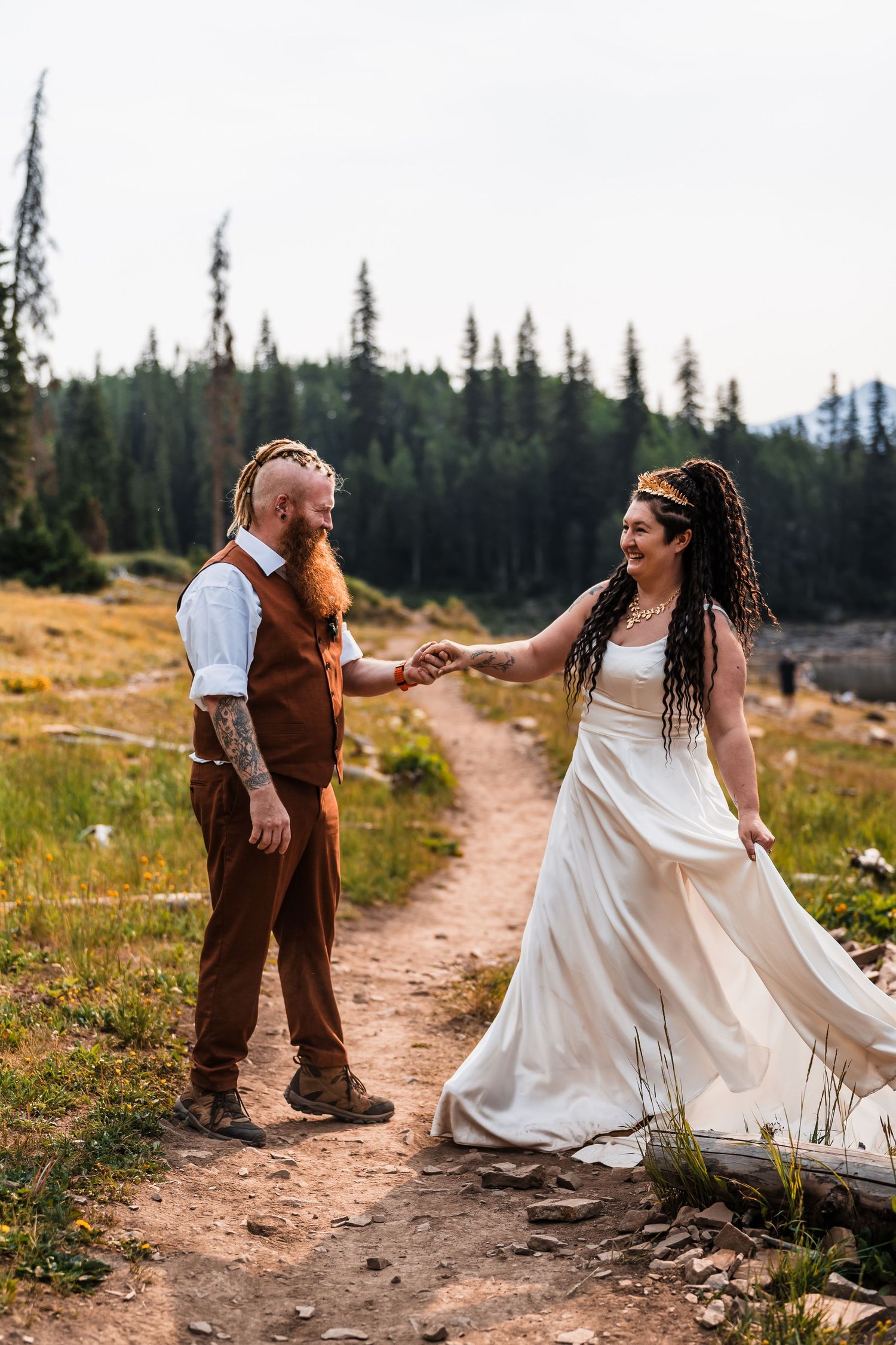 Bohemian bride in white dress and groom in brown vest hold hands on forest trail at Colorado mountain elopement.