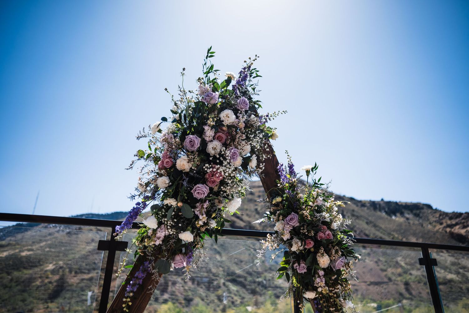 Floral wedding arch adorned with pink and purple blooms against a clear blue sky and mountain backdrop.