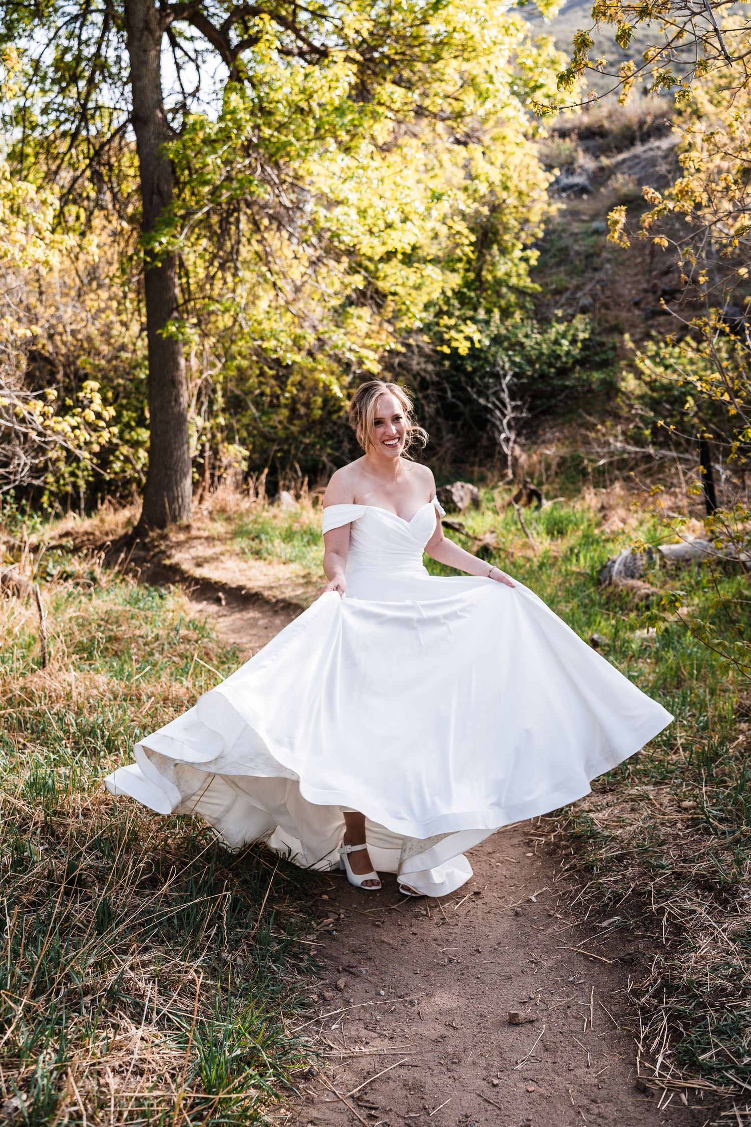 Bride in white off-shoulder gown twirling on a forest path surrounded by autumn trees.