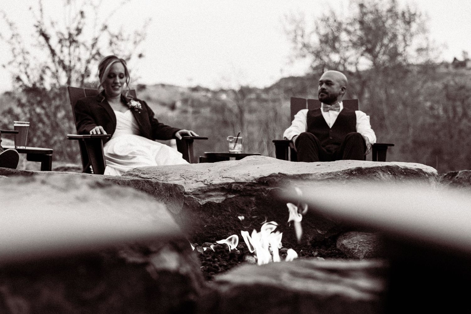 Black and white photo of bride and groom relaxing by a stone fire pit outdoors at their rustic wedding.