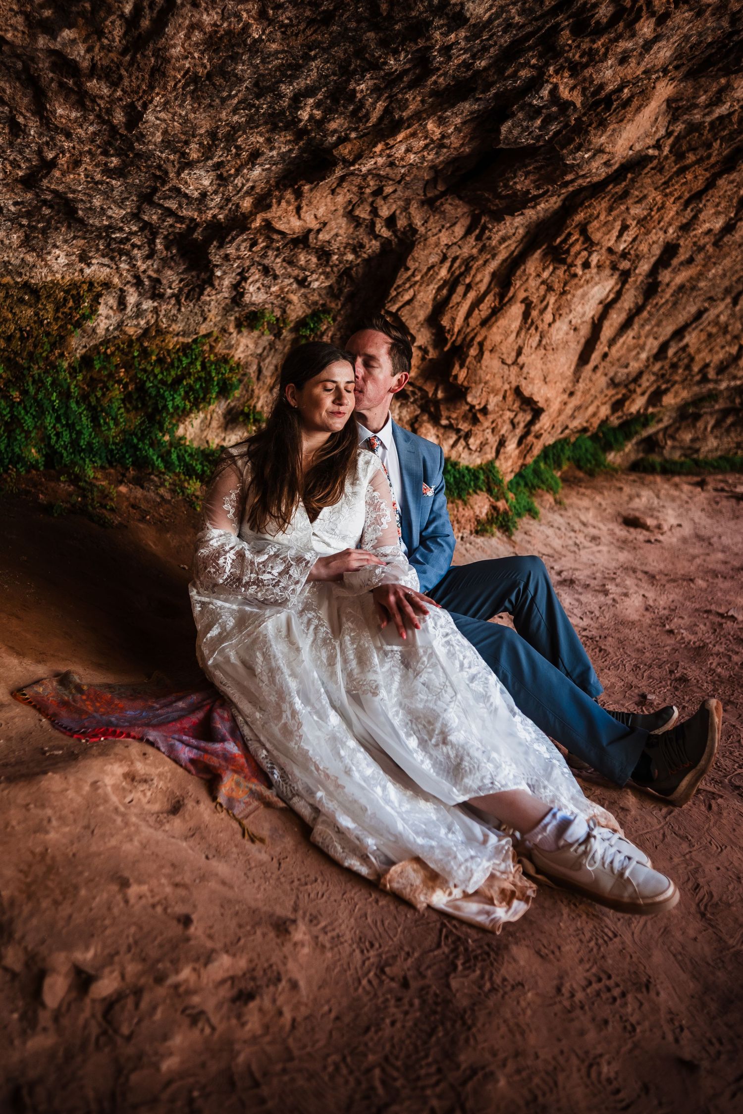 Bride and groom sitting together under a rocky cave overhang on red sand during elopement.
