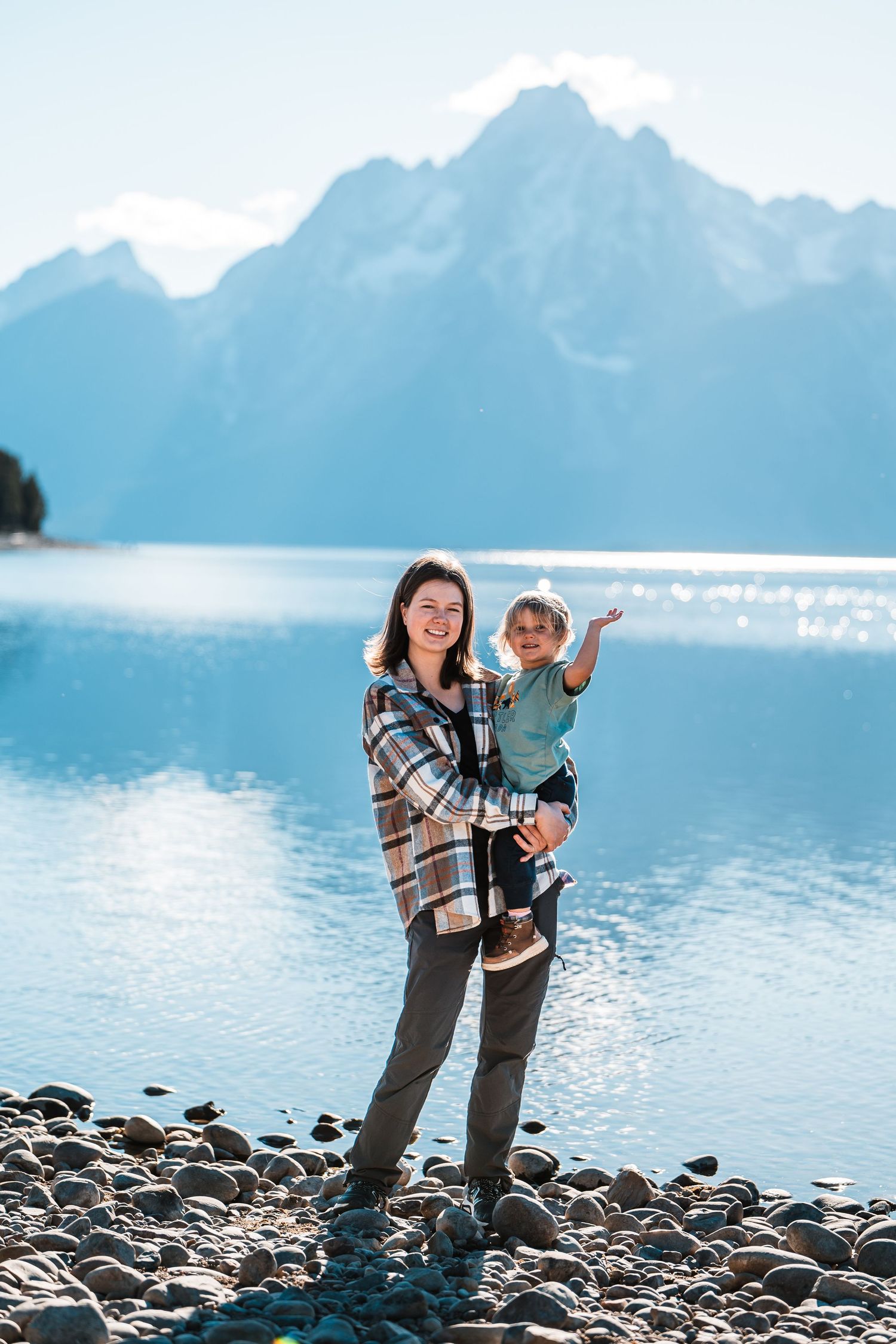 A parent and child share a playful moment on a rocky lakeshore with snow-capped mountains in the background.