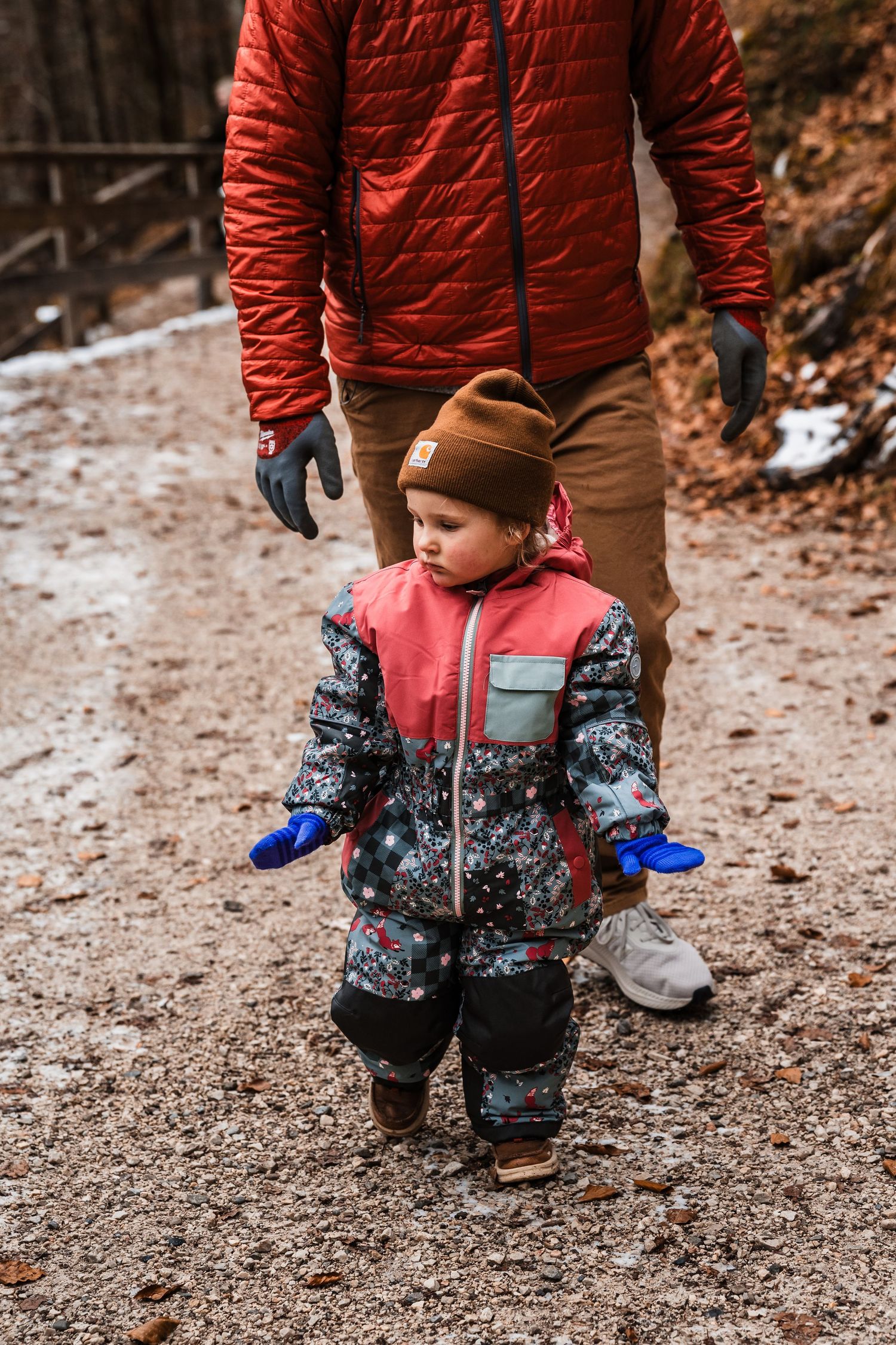 Child wearing red and patterned winter coat stands on snowy forest trail with adult nearby.