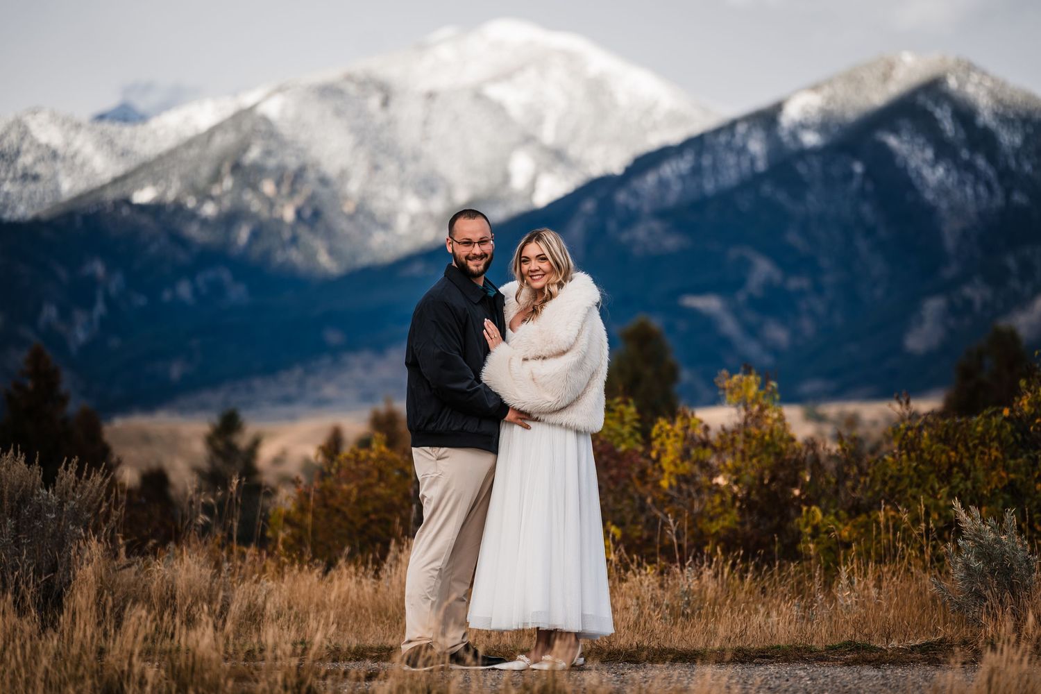 A couple embraces against a backdrop of snow-capped mountains and autumn foliage in a romantic outdoor setting.