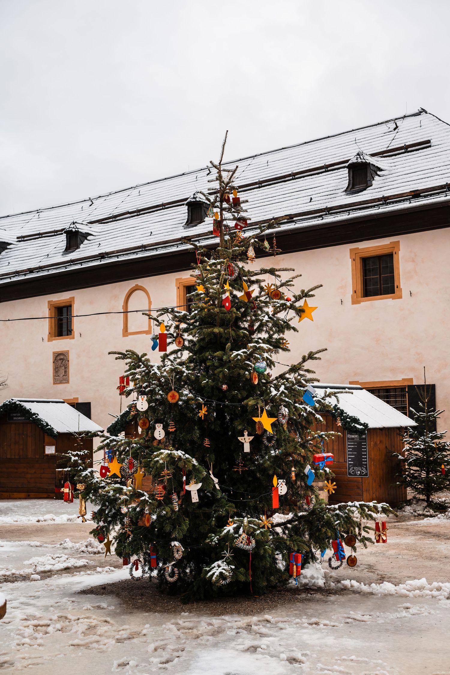 A decorated Christmas tree stands in the snow against a white building with wooden doors and windows.
