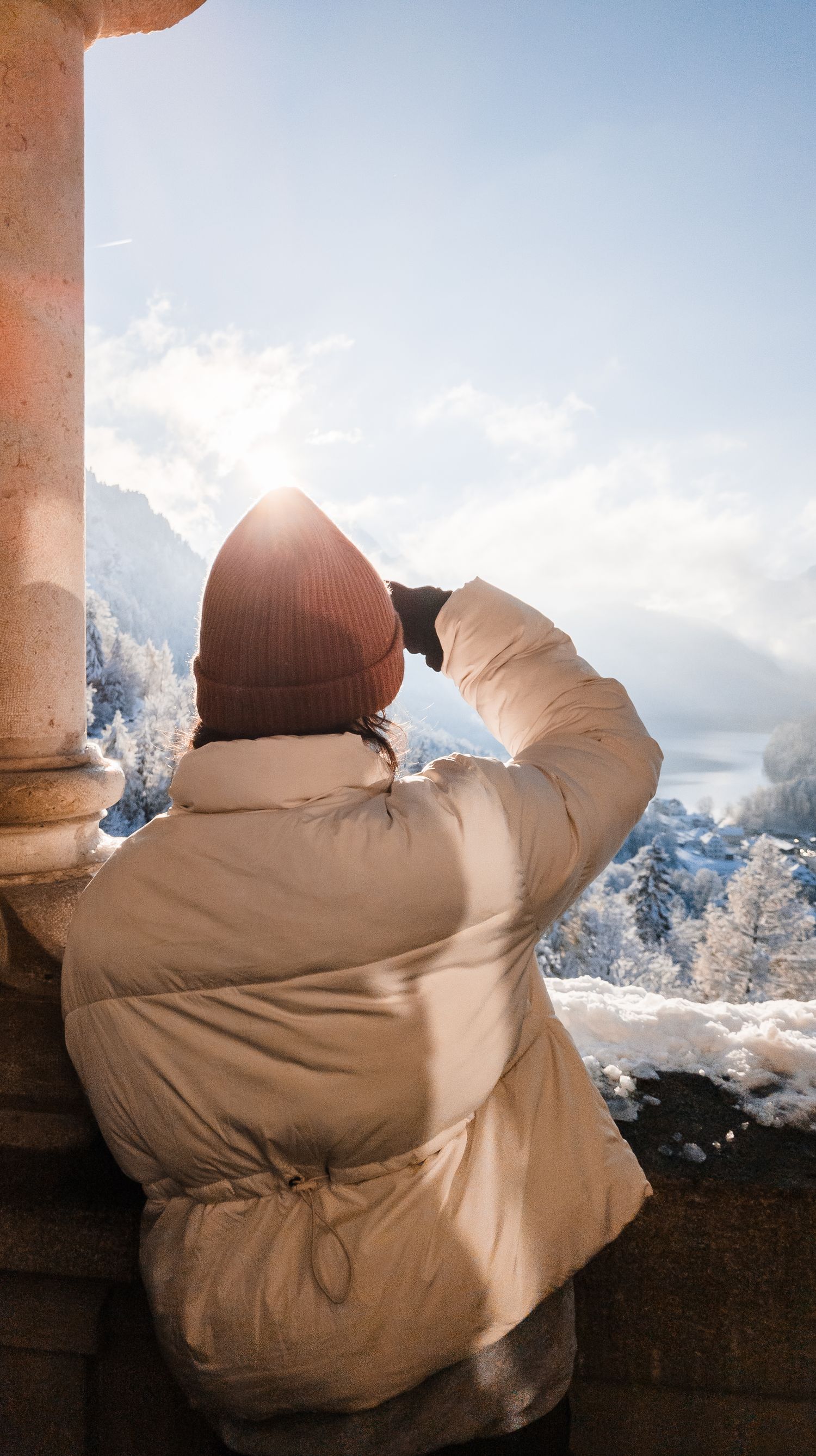 Person wearing beanie and beige jacket photographs snowy mountain landscape from scenic viewpoint.