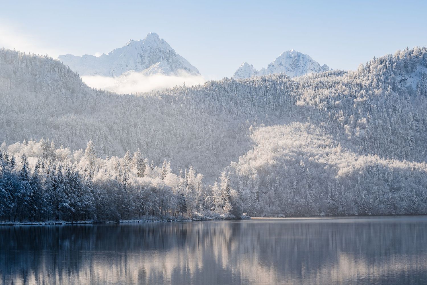 Snow-capped mountain peaks reflect in a pristine alpine lake surrounded by evergreen forest on a winter morning.
