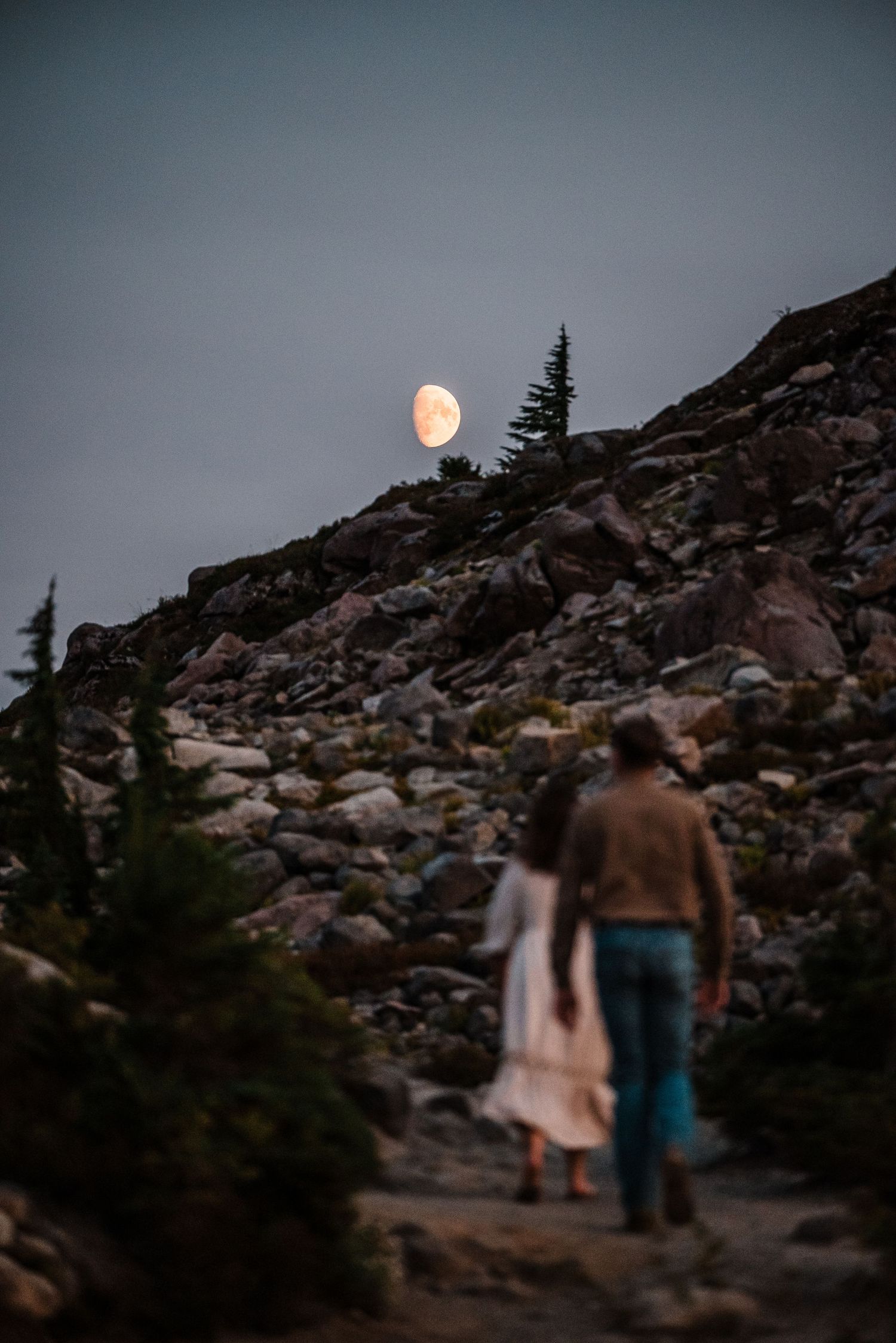 Full moon rises over rocky mountain terrain at dusk with figures walking in silhouette.