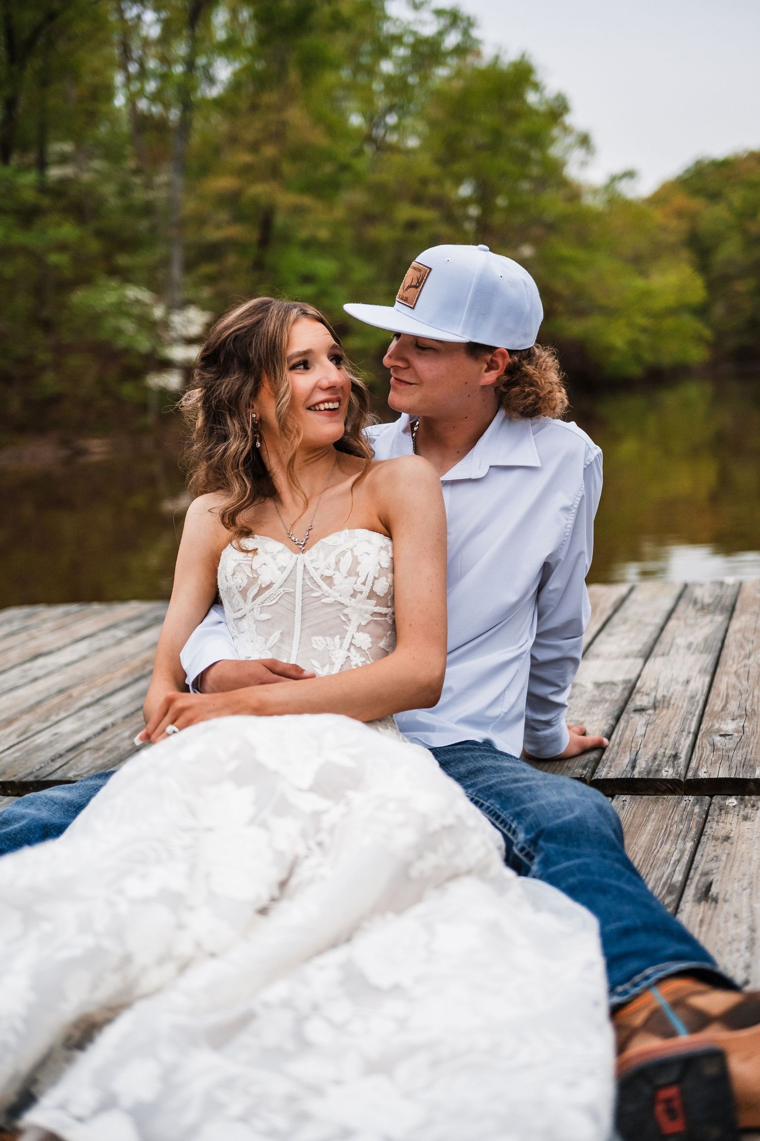 Romantic couple sitting on wooden dock by lake with bride wearing white lace dress.