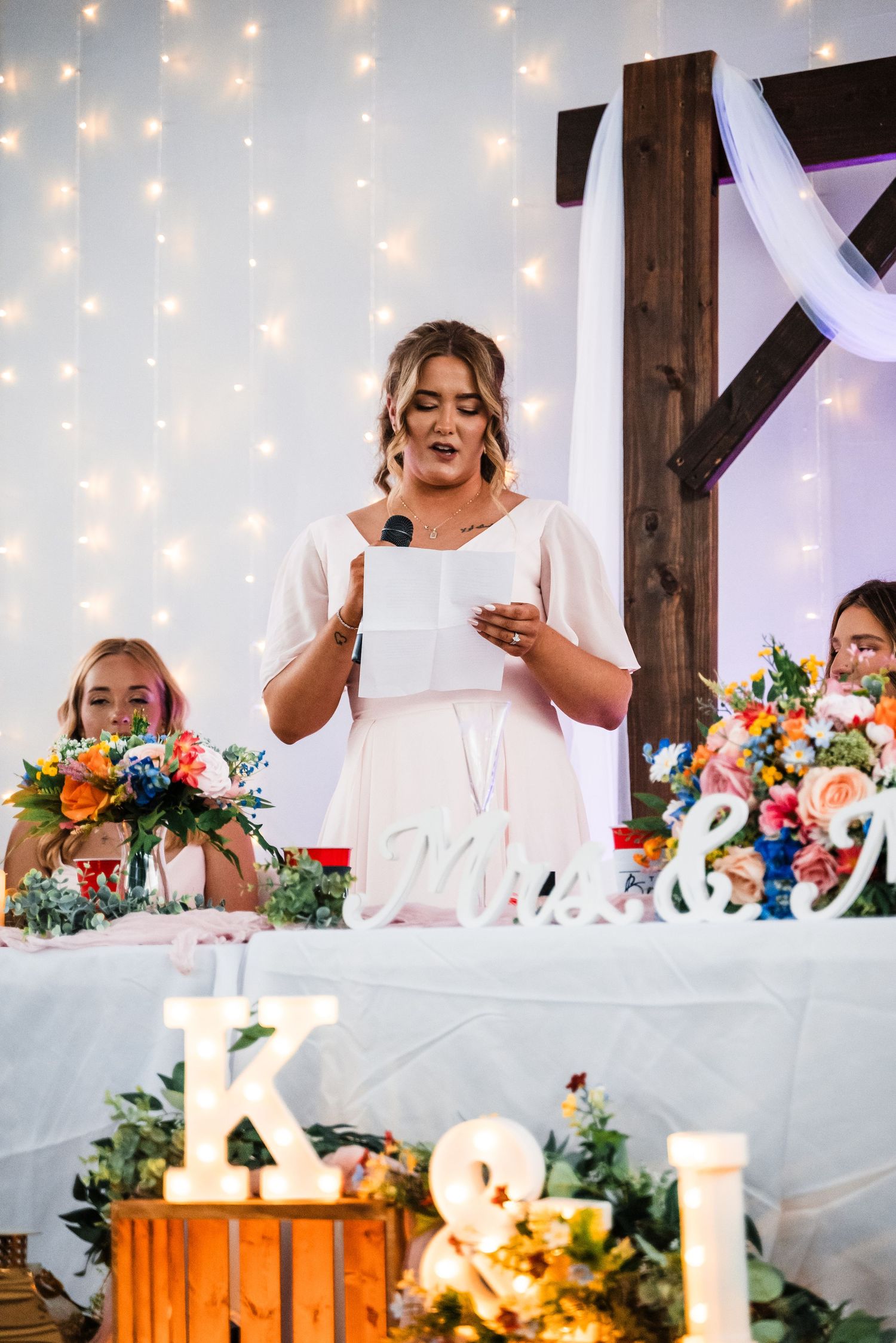 Speech being delivered at wedding reception with decorative lights and floral arrangements on head table.